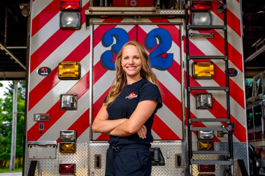 Woman in firefighter uniform standing in front of fire truck with arms crossed, smiling outdoors.