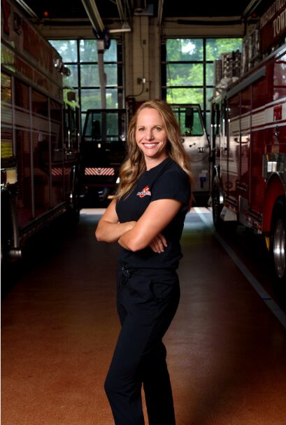 Young woman with long hair smiling, standing with arms crossed between fire trucks inside a fire station, large windows behind.