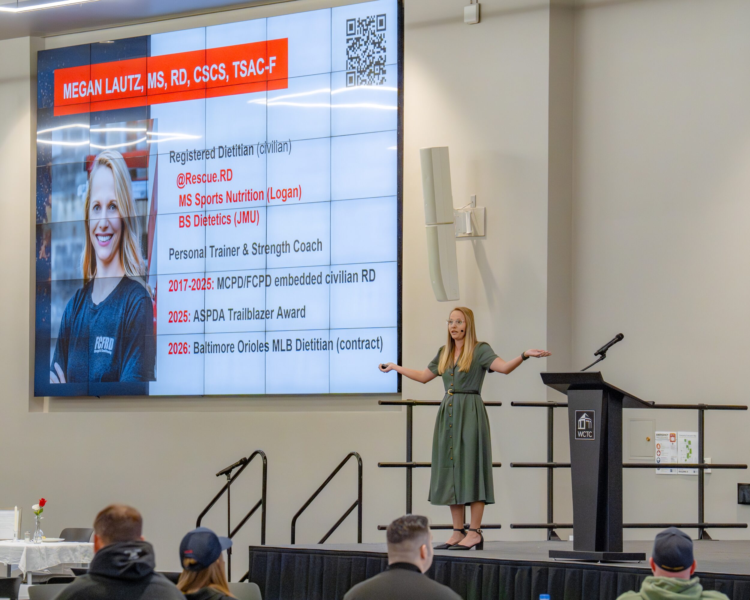 Woman in green dress speaking on stage with a large screen displaying her credentials and information behind her.