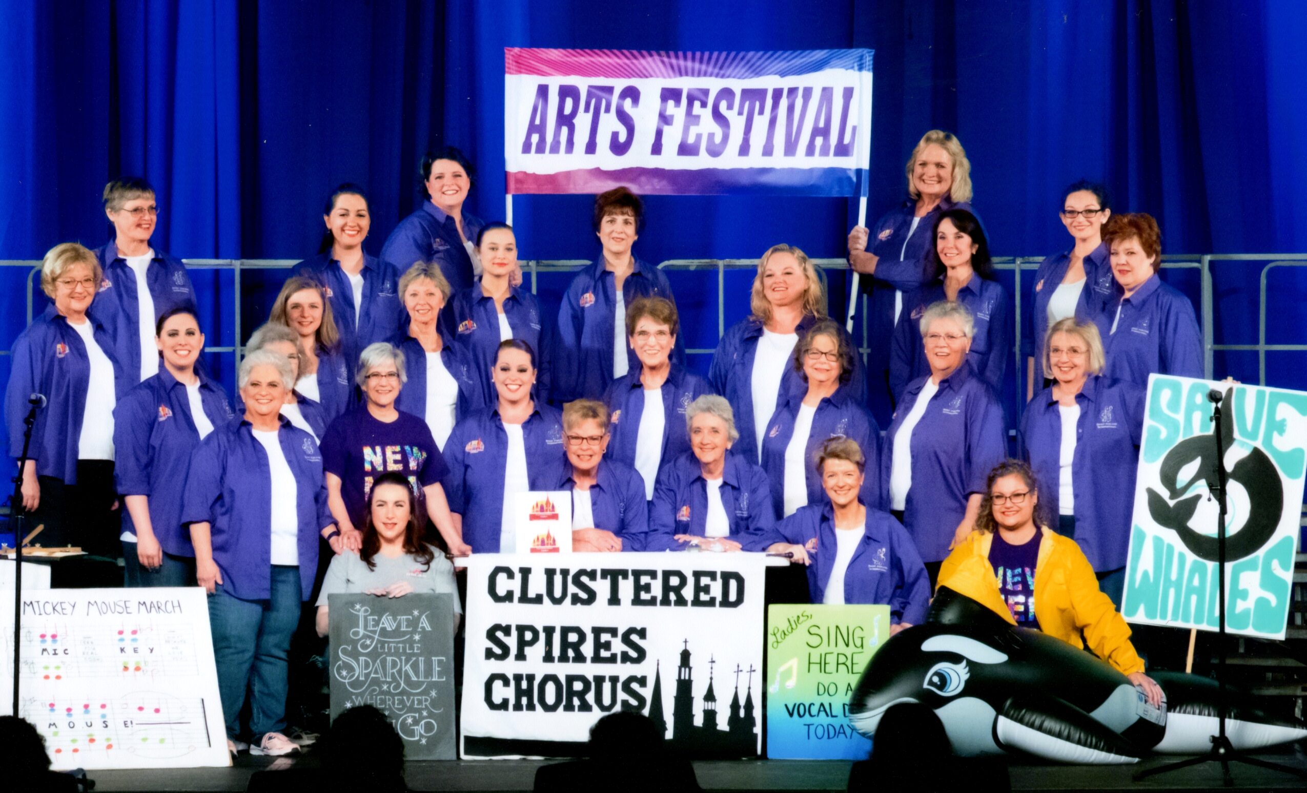 Group of people on stage with a banner reading 'Arts Festival,' holding signs and wearing blue jackets.