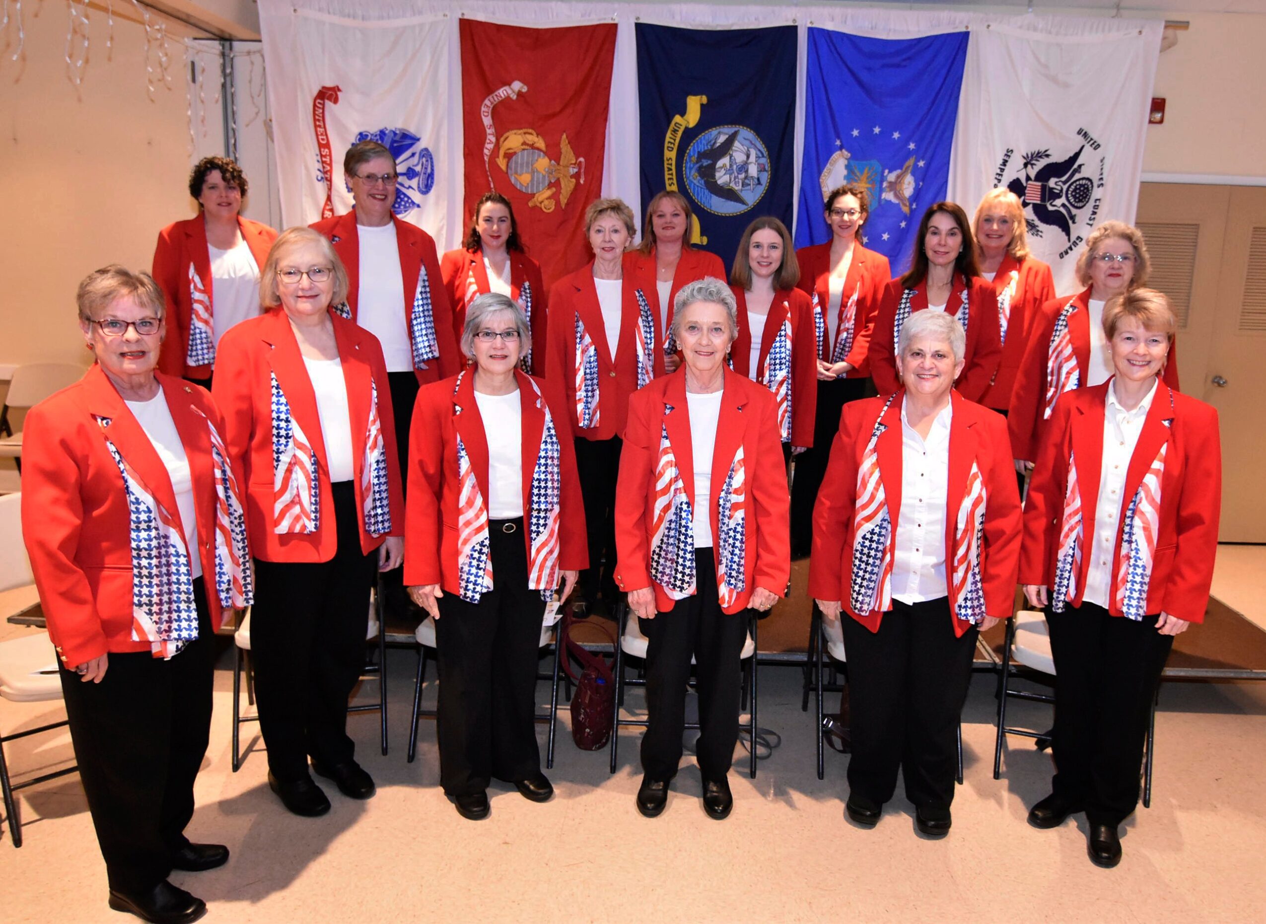 Group of 15 women in red jackets and white shirts standing in front of colorful flags, smiling indoors.