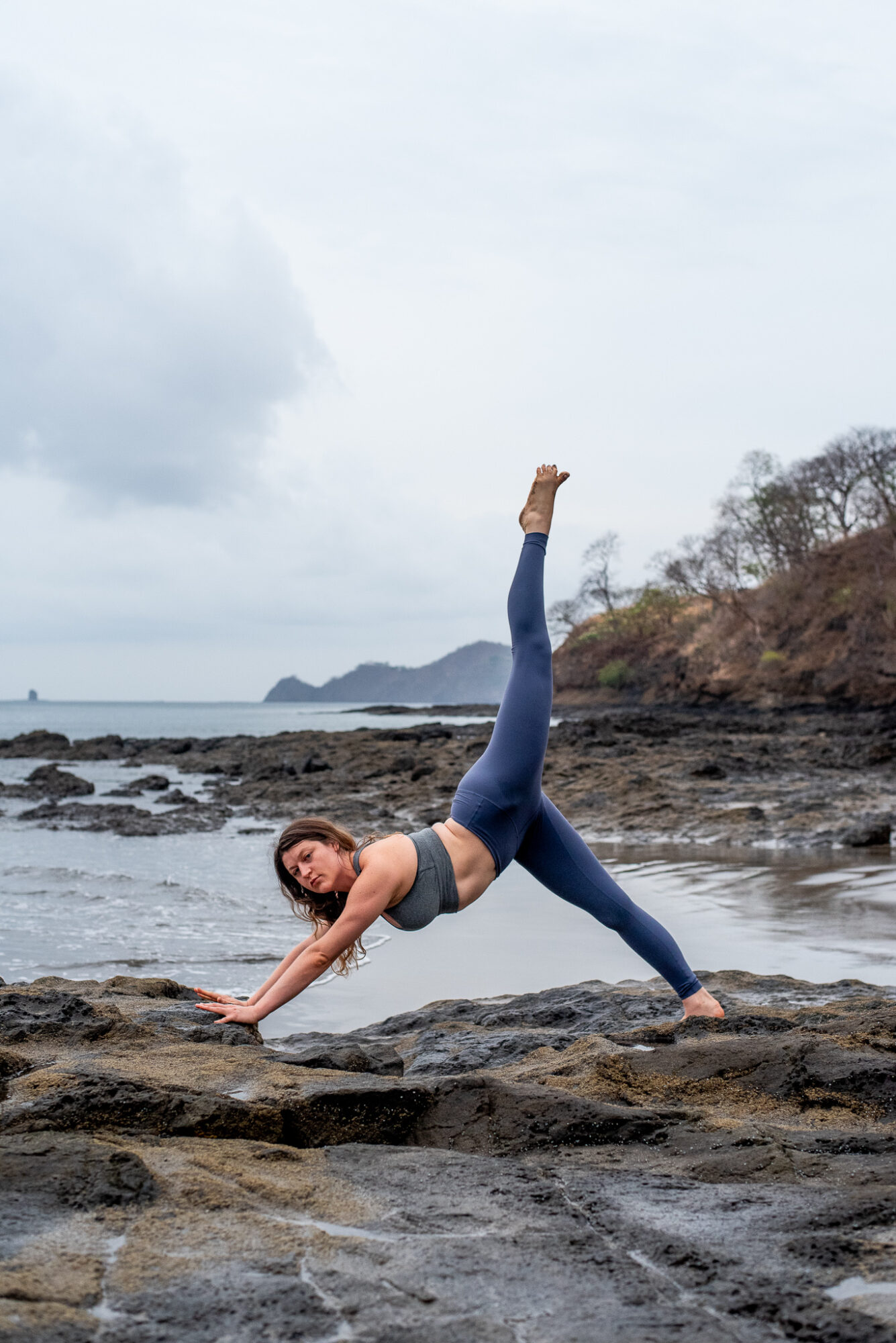 Person performing a yoga pose on rocks by the water, with one leg raised and arm extended on the ground.