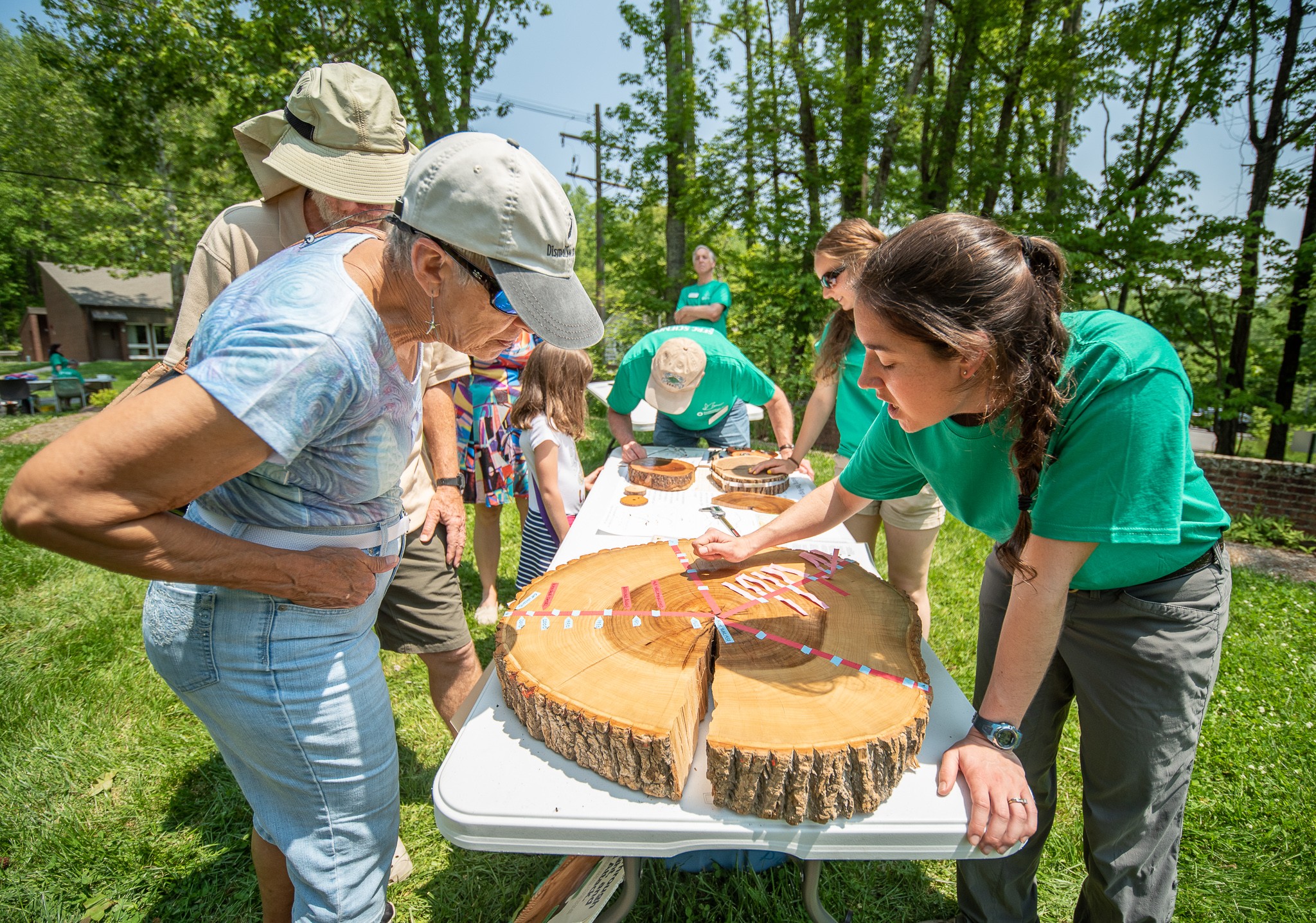 People gather around a table with a large cake outdoors, trees and blue sky in the background.