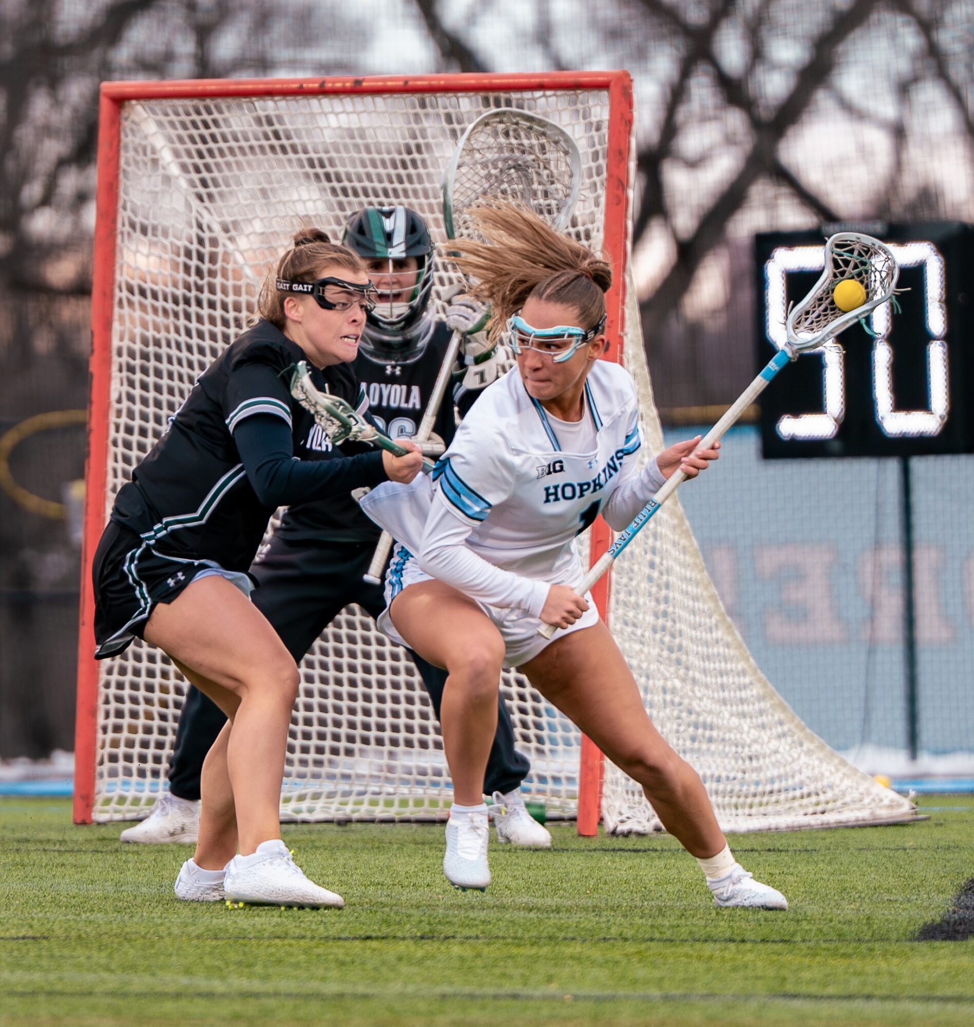 Lacrosse players and goalie in action on field, with goal and scoreboard in background.