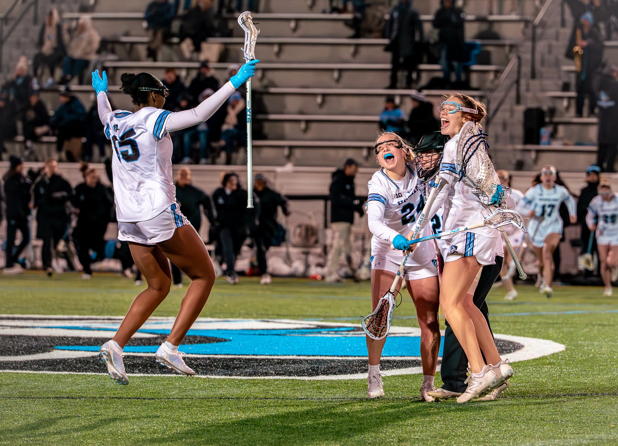 Group of female lacrosse players celebrating on field, with one player raising arms, others smiling and holding sticks, spectators in background.