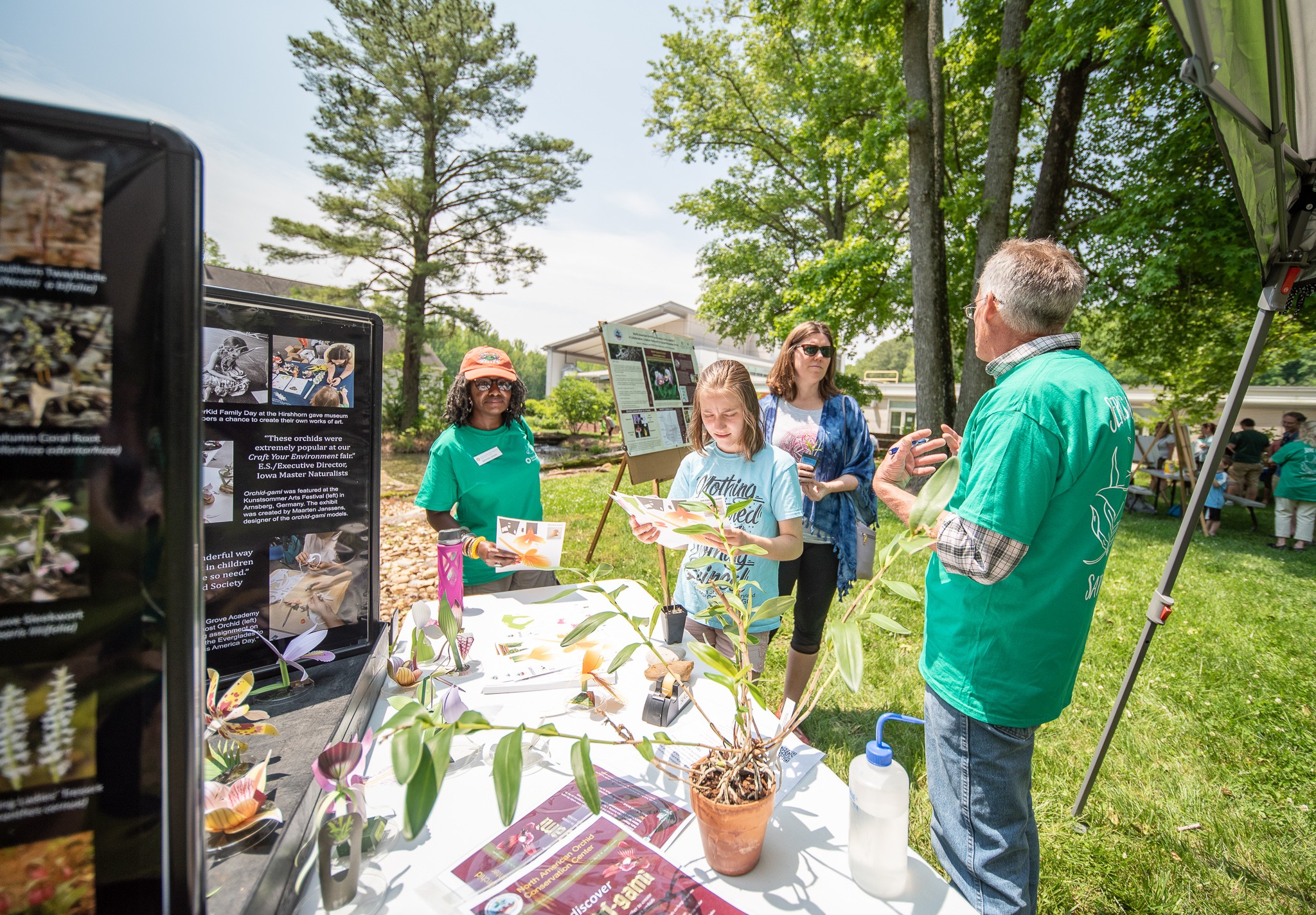 People gather outdoors around informational display tables with trees and grass in the background.