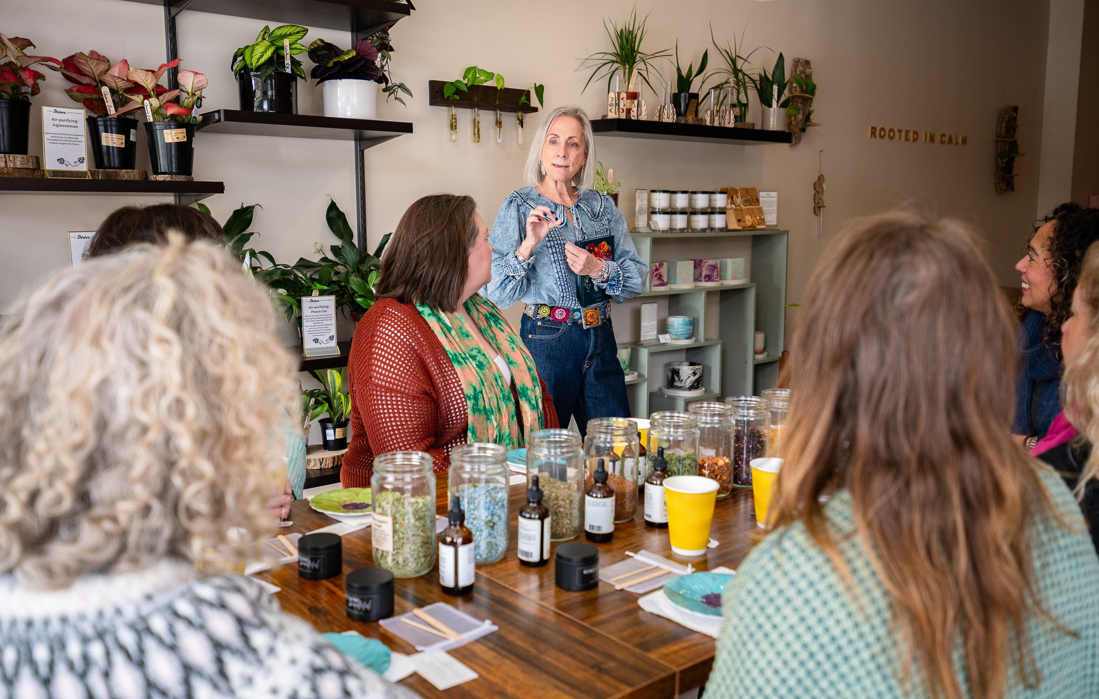 Group of women seated around a table listening to a woman speaking in a room with plants and shelves.