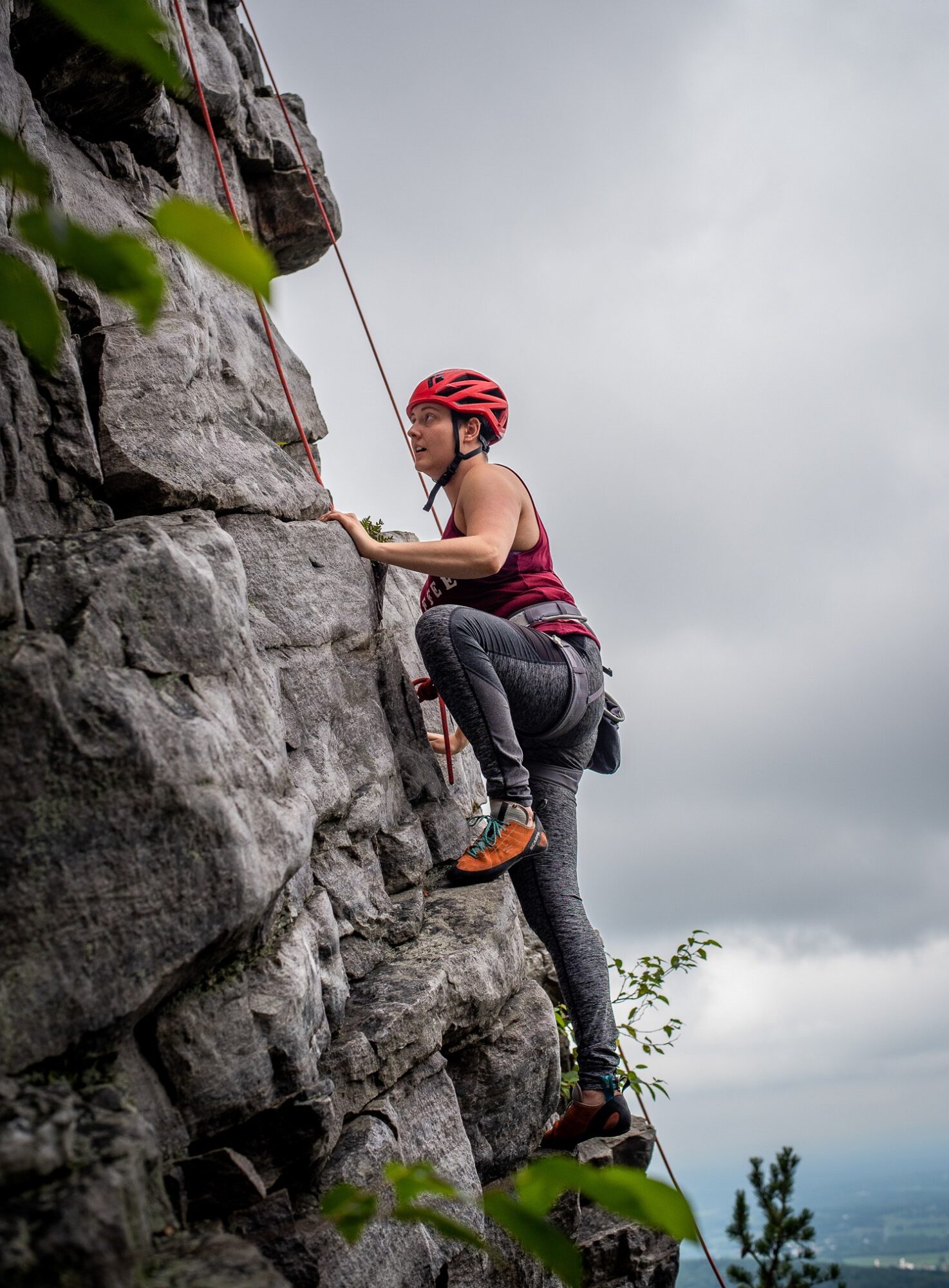 Person climbing a rock wall outdoors, wearing a red helmet and climbing gear, with cloudy sky background.