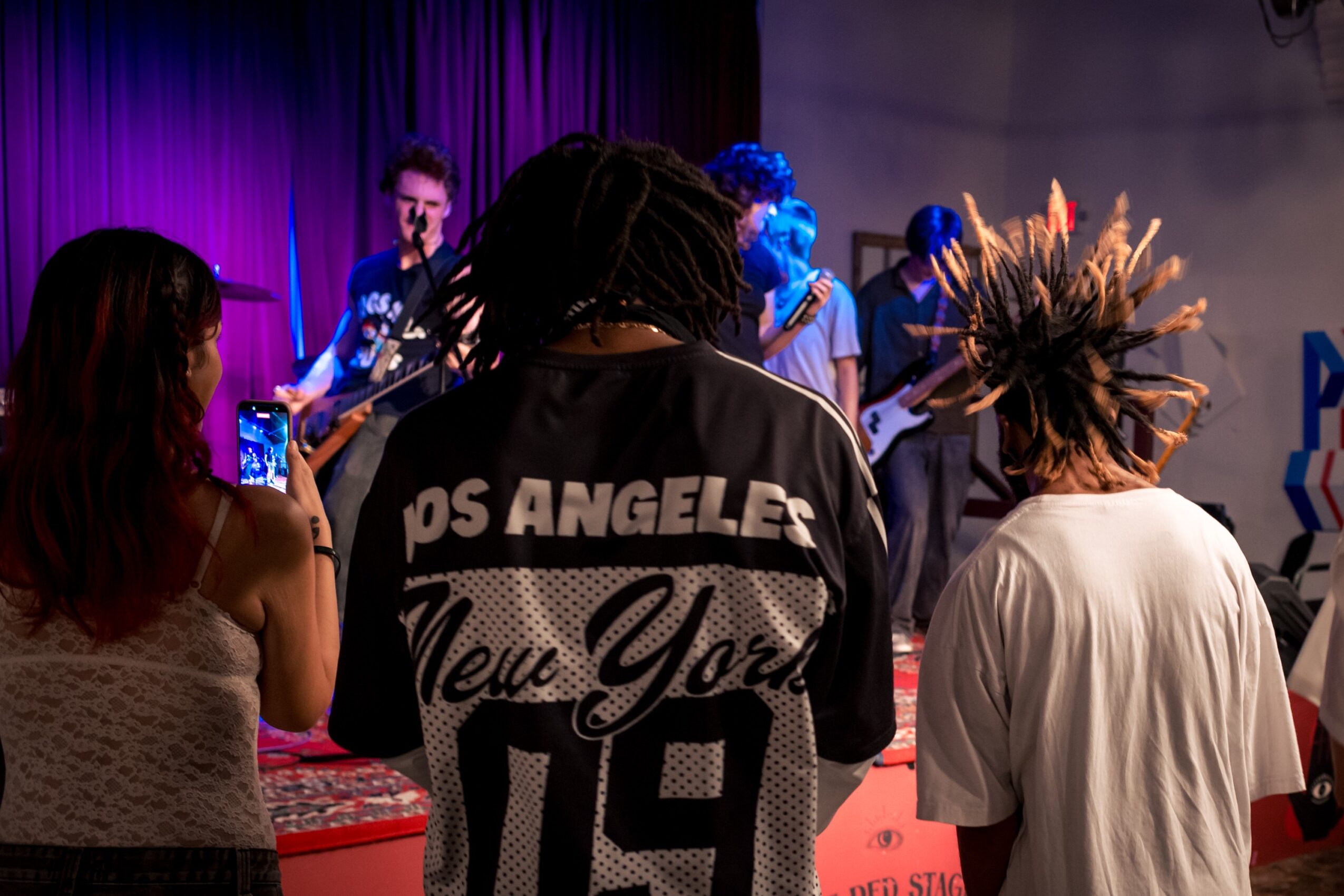 People watching a band perform on stage, one person taking a photo, stage with purple lighting, and a person with spiky hair.