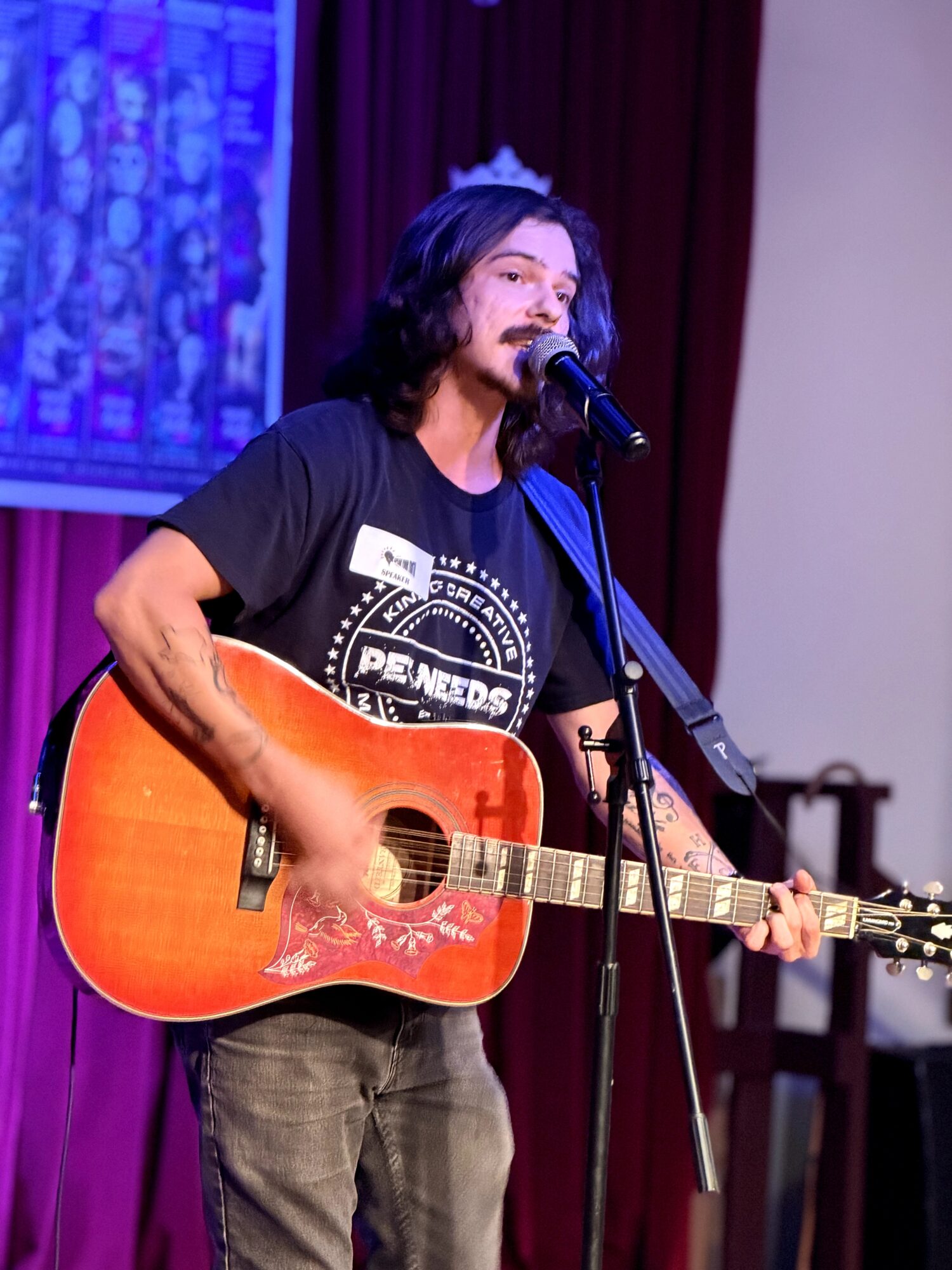 Person with long dark hair singing into a microphone while playing an orange acoustic guitar, wearing a black T-shirt.