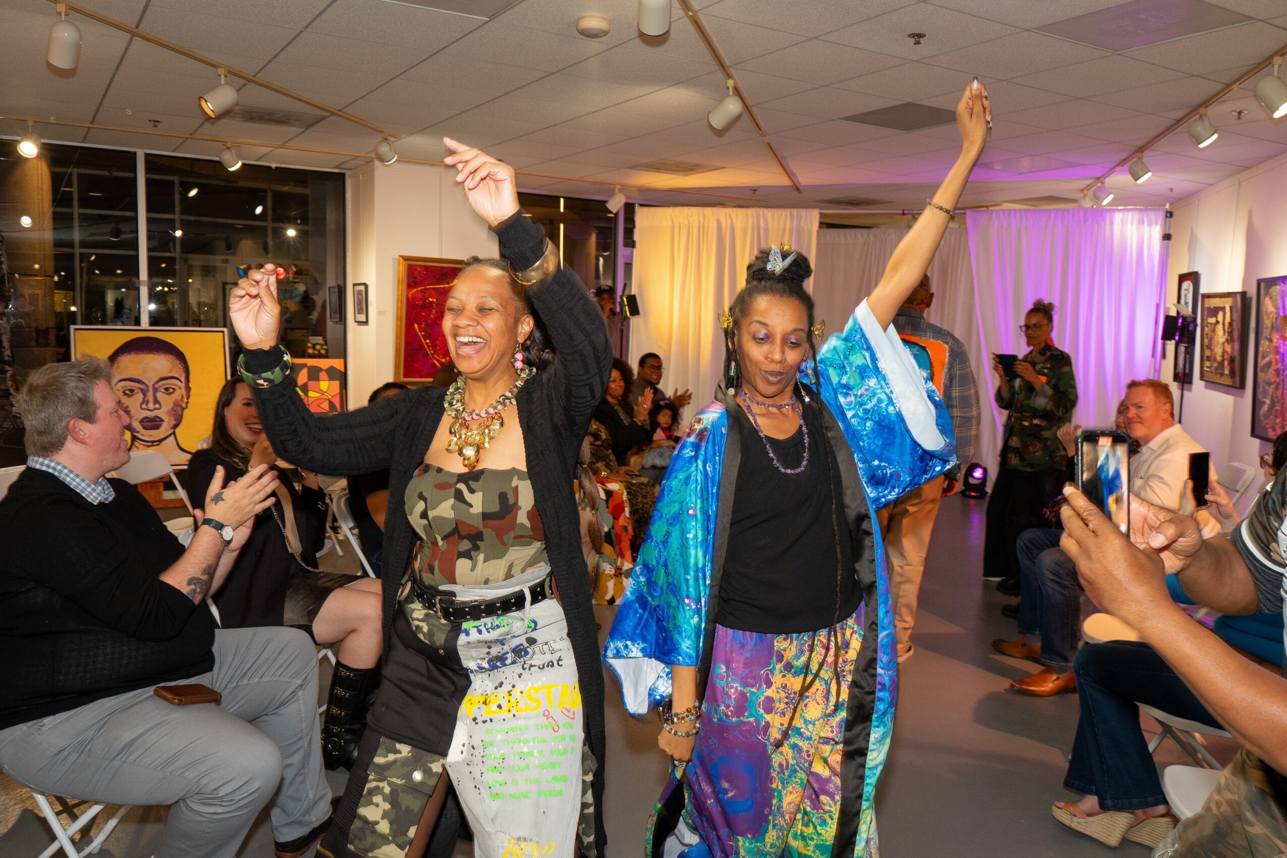 Two women dancing and celebrating at an indoor event, with people clapping and taking photos in the background.