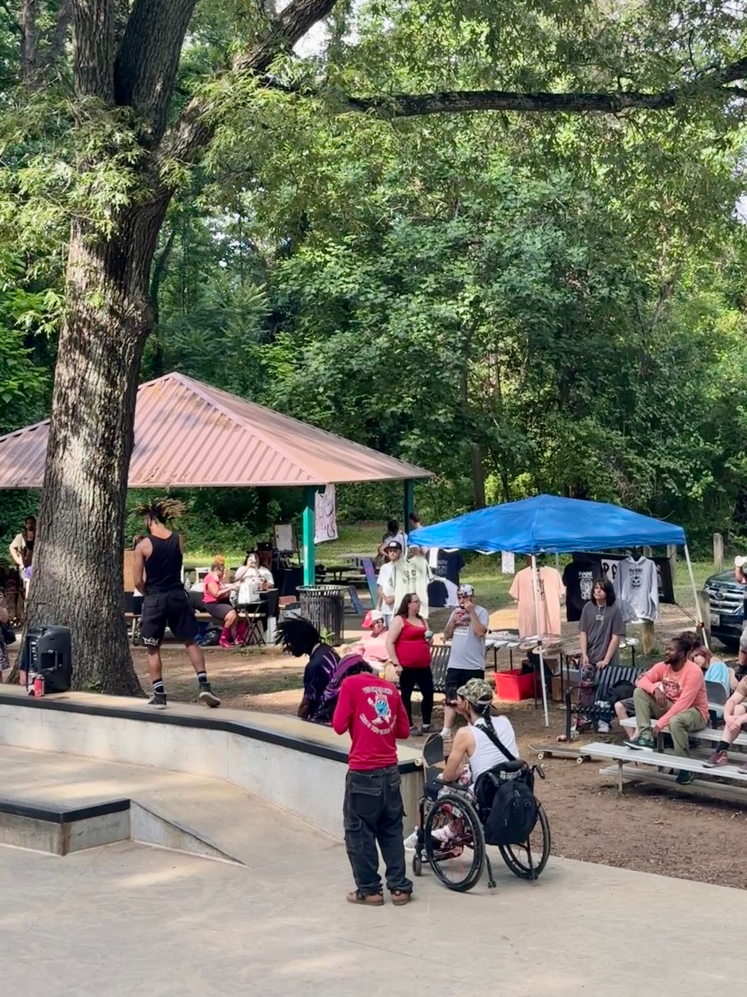 People gather in a park with trees, a pavilion, and a blue canopy, some sitting and others standing, with a person in a wheelchair.