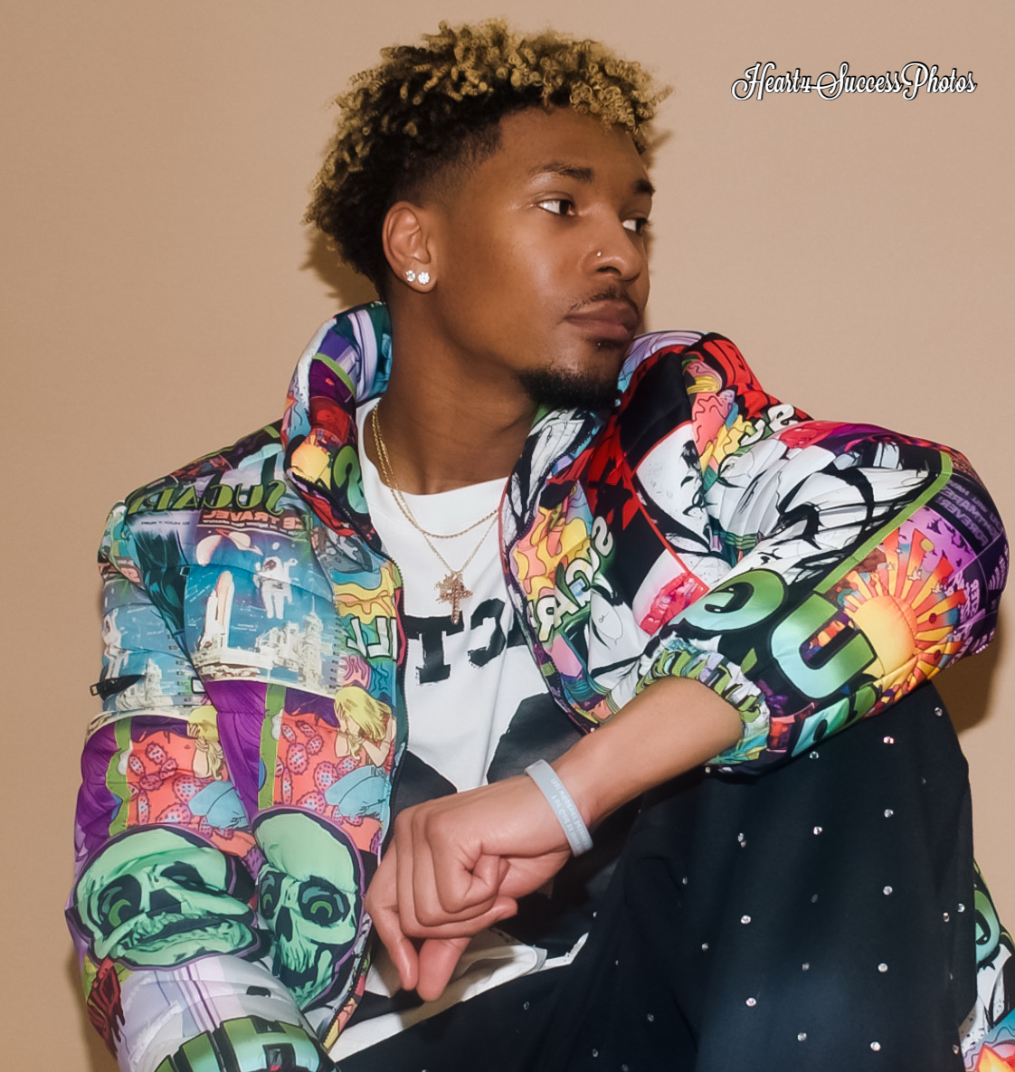 Young person with curly hair wearing a colorful jacket, sitting with arm resting on knee, against a plain background.