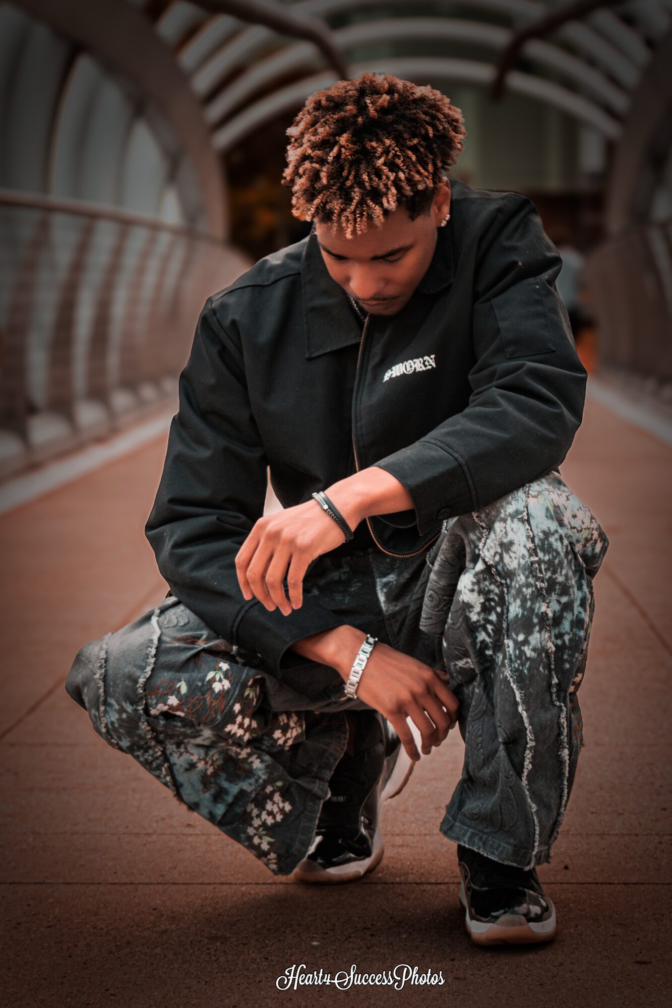 Person crouching on a bridge with metal railings, wearing a black jacket and patterned pants, looking down.
