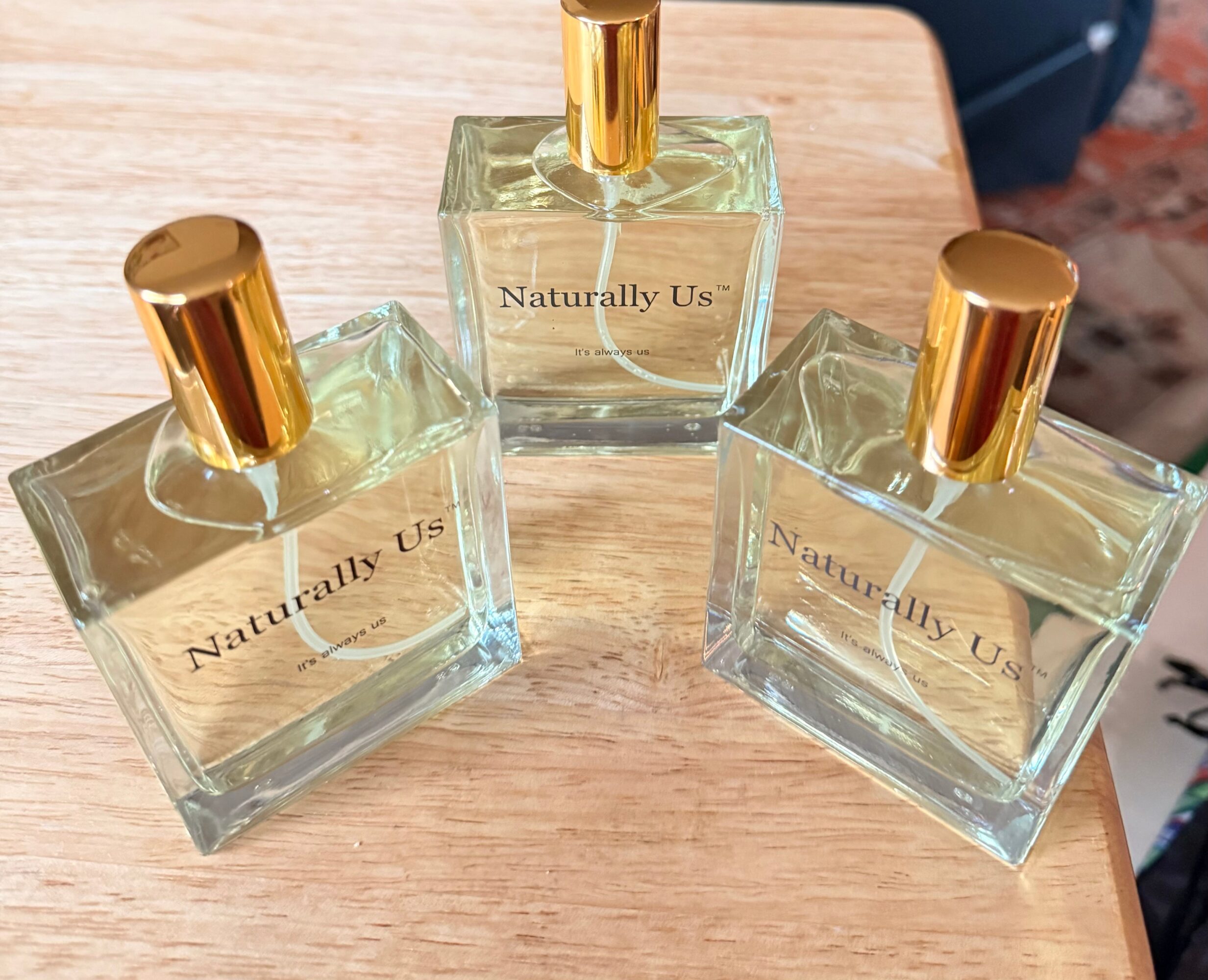 Three glass perfume bottles with gold caps on a wooden surface, labeled 'Naturally Us'.