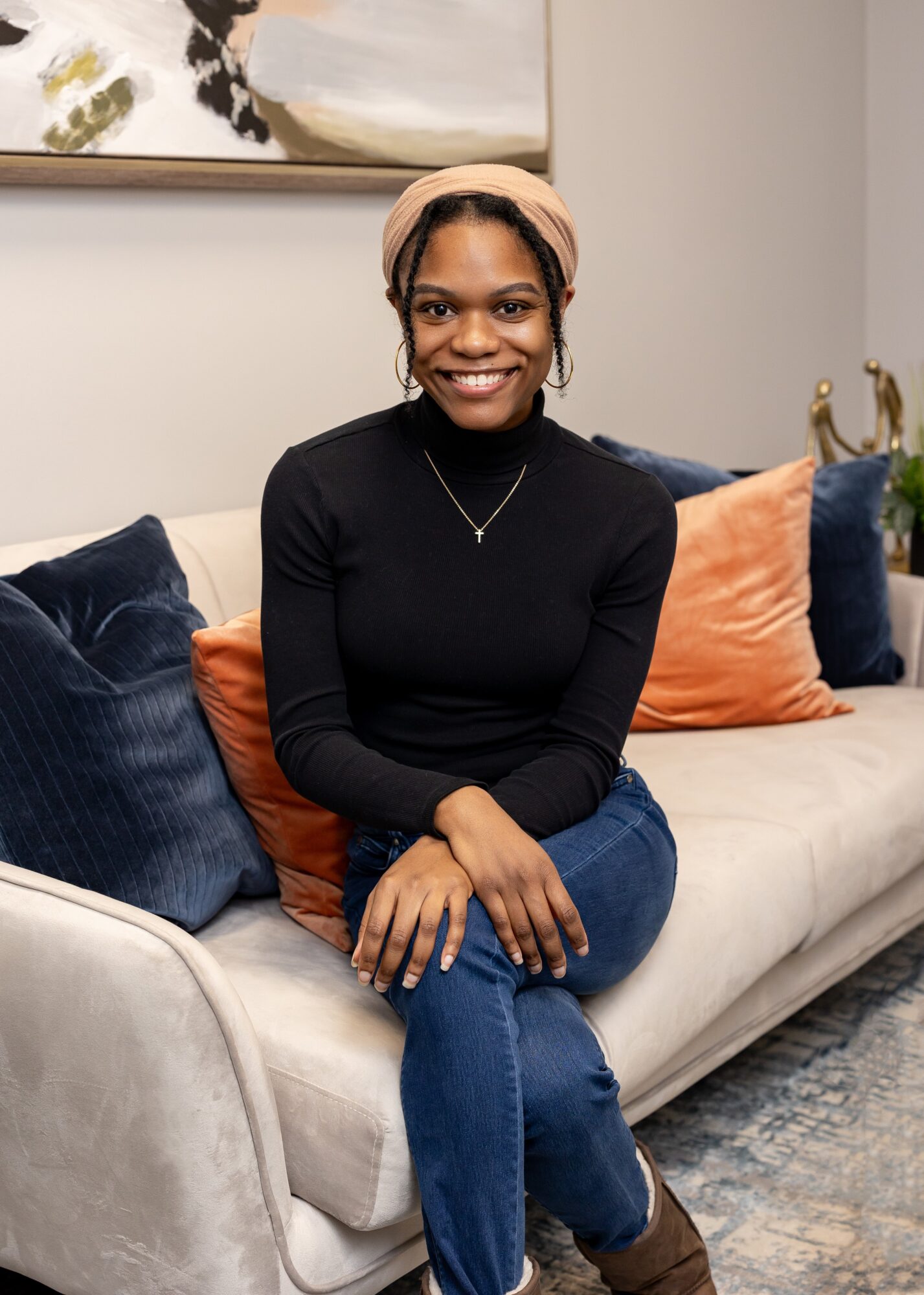 Smiling woman sitting on a sofa with colorful pillows, wearing a black top and beige headband.