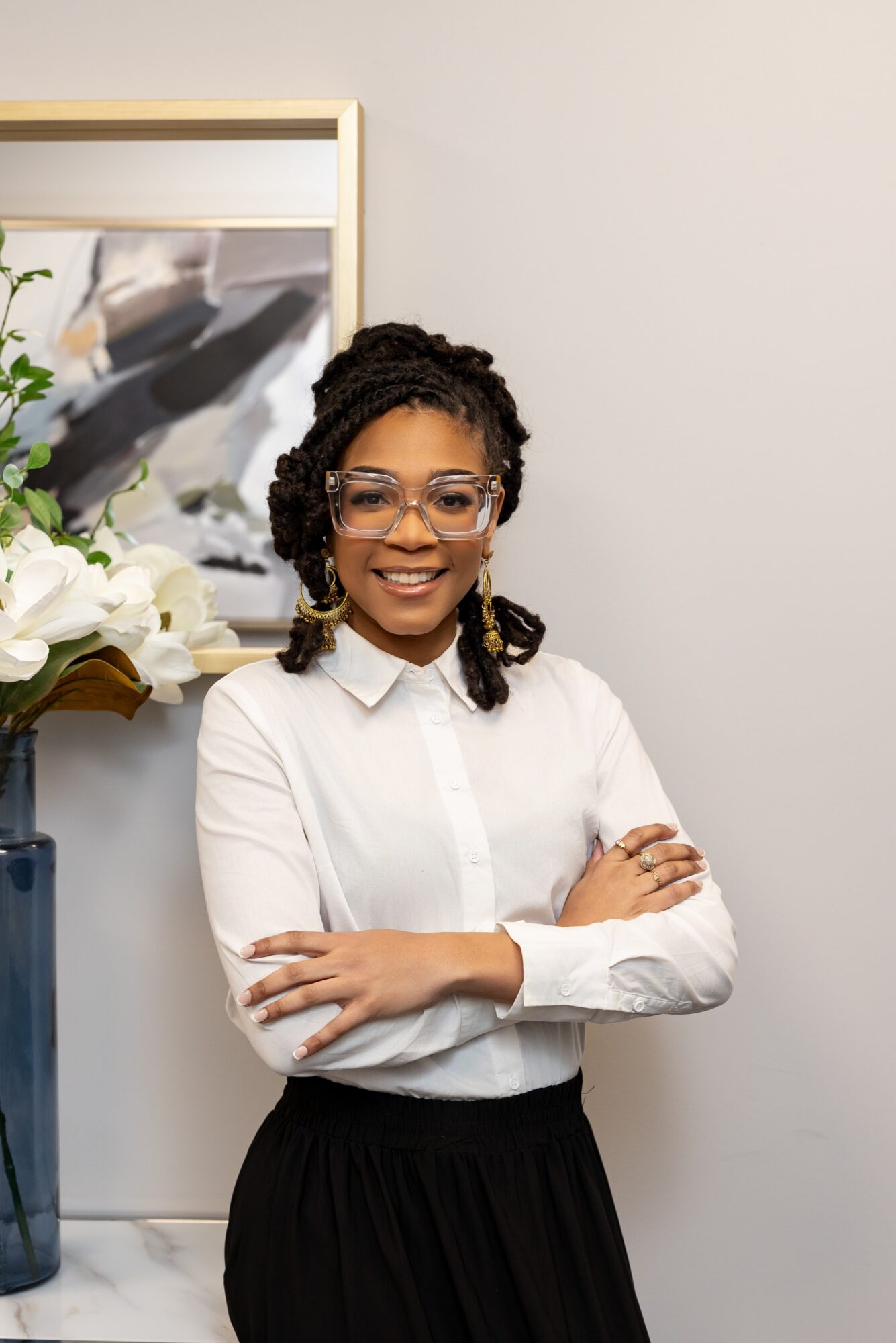 Smiling woman with glasses and braided hair stands with arms crossed in a white shirt and black skirt, in a room with a mirror and flowers.