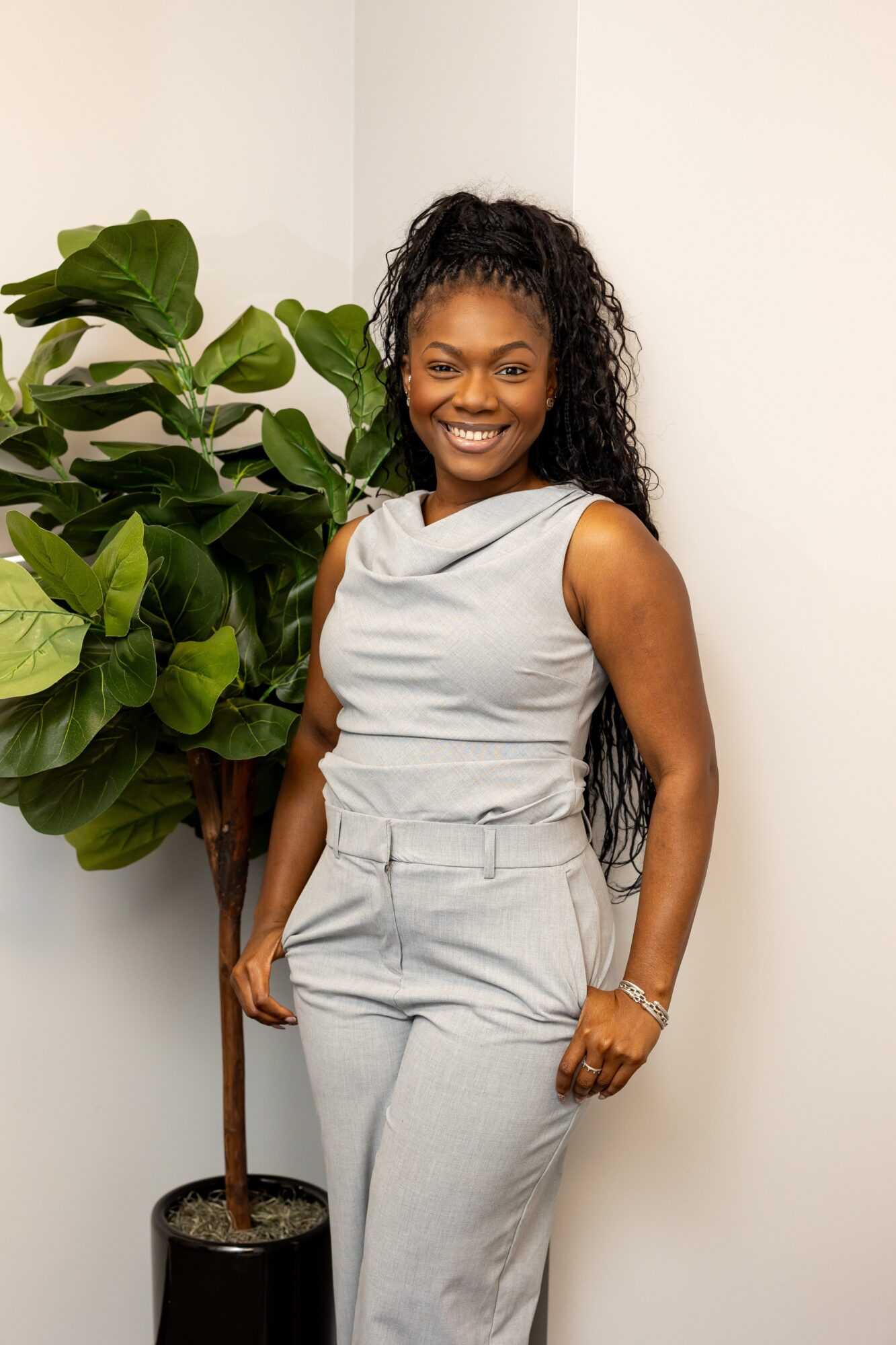 Smiling woman with long curly hair stands next to a large potted plant, wearing a sleeveless top and high-waisted pants.