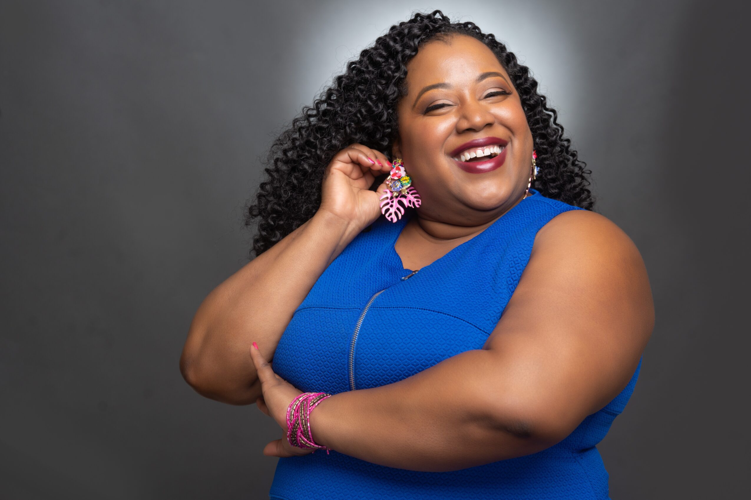 Smiling woman with curly hair wearing a blue dress and pink jewelry, posing with her hand near her ear.