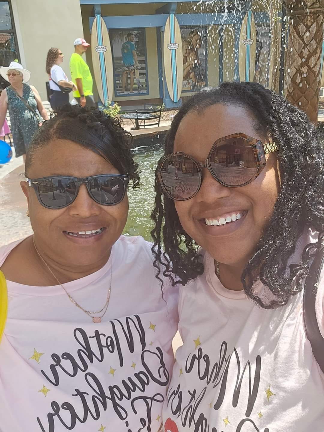 Two women smiling outdoors, wearing sunglasses and white shirts with black text, with a colorful background and other people nearby.