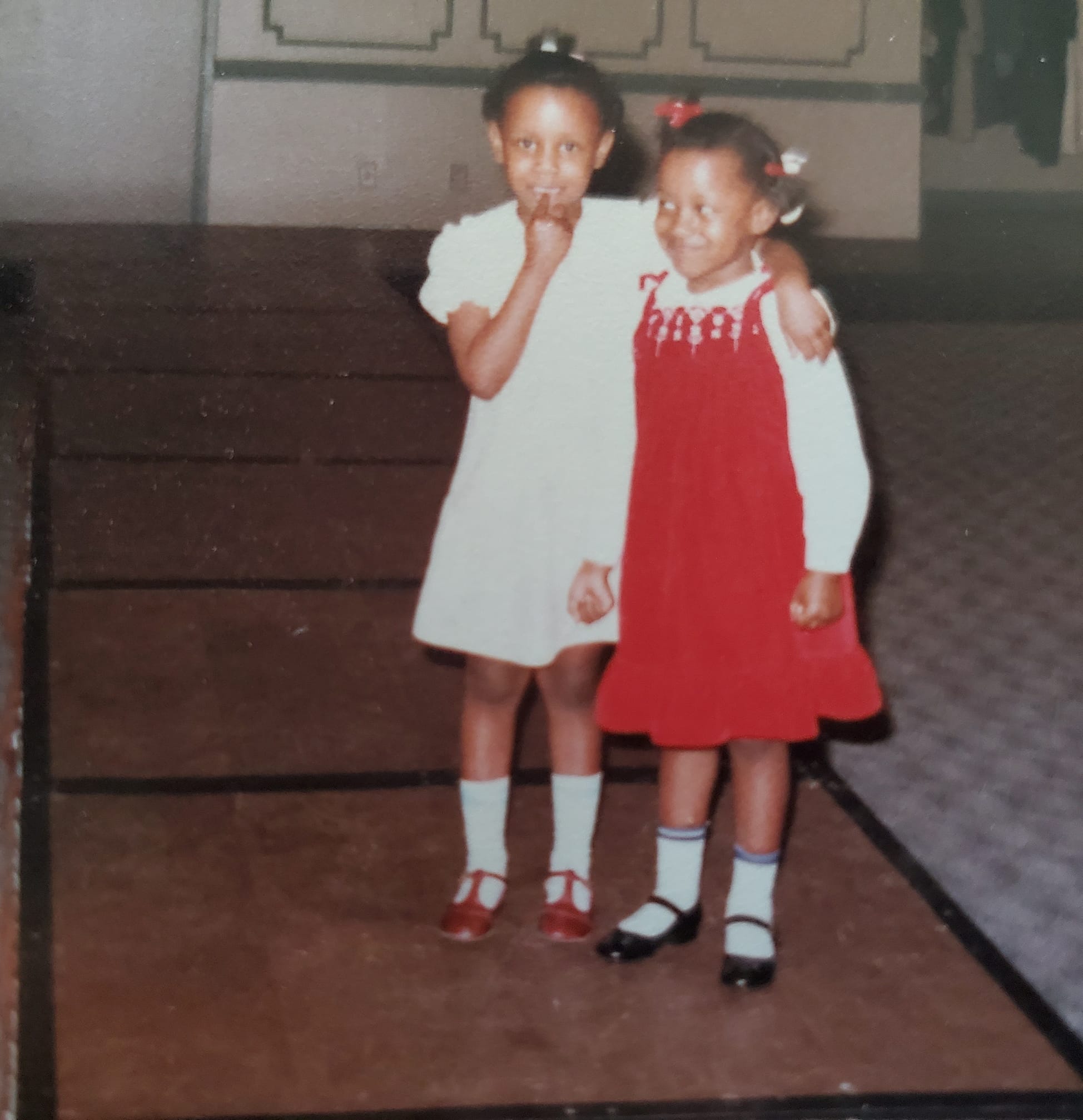 Two young girls standing together indoors, one in a white dress and the other in a red dress, smiling.