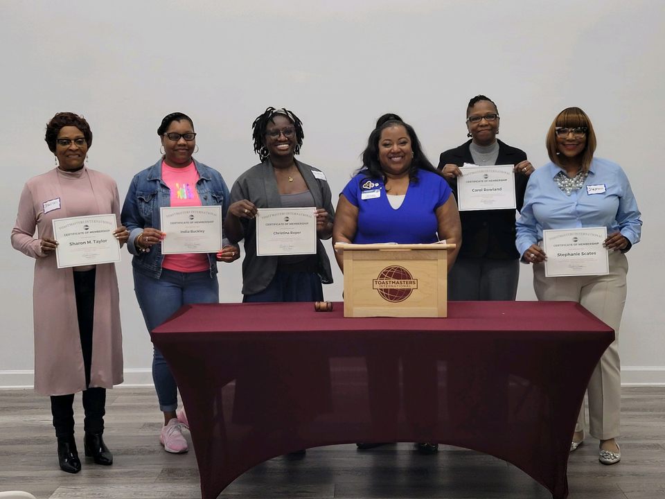 Seven people standing behind a table holding certificates, with a box on the table, in a room with a plain background.