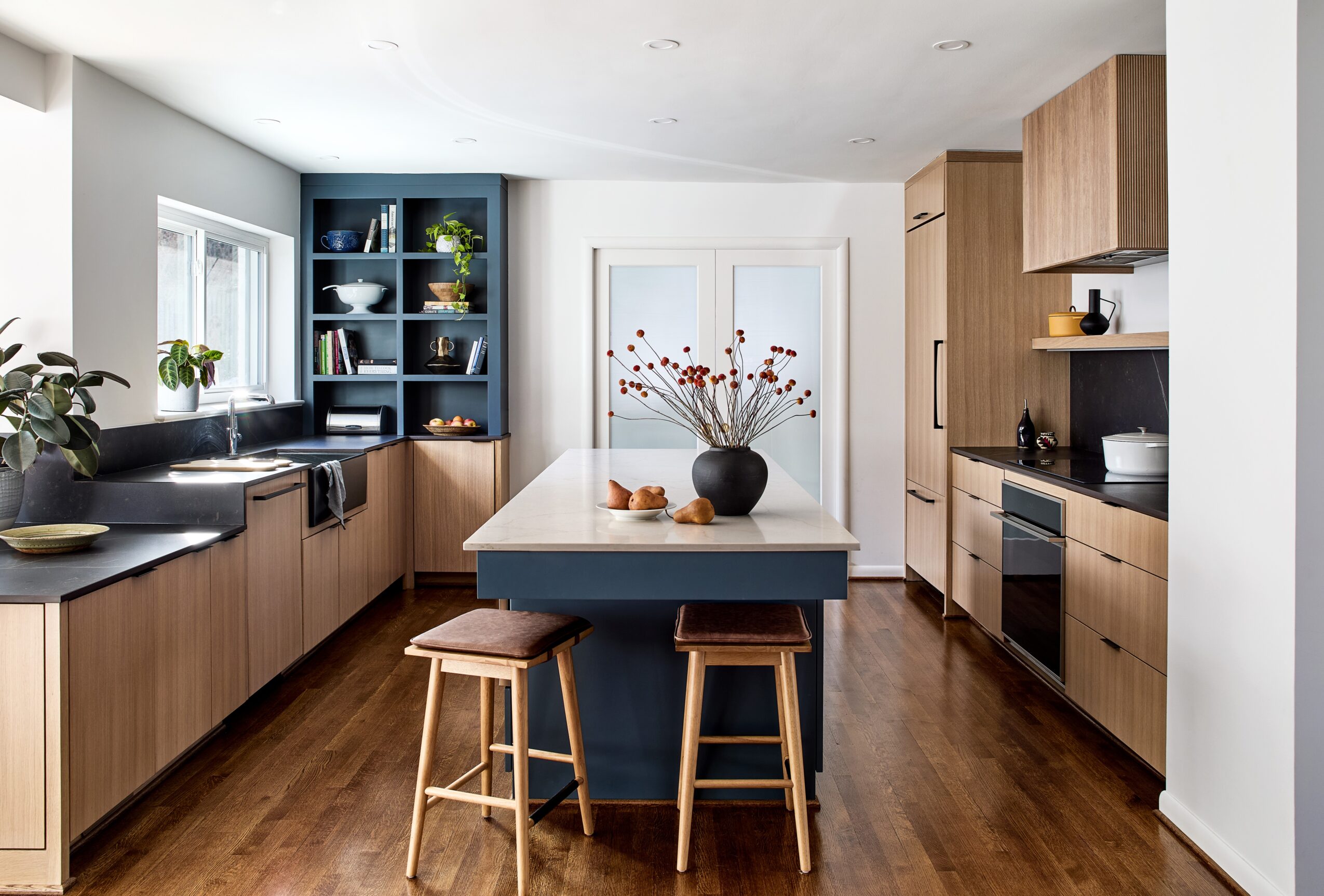 Modern kitchen with wooden cabinets, a central island with stools, and a window with plants and decorative items.