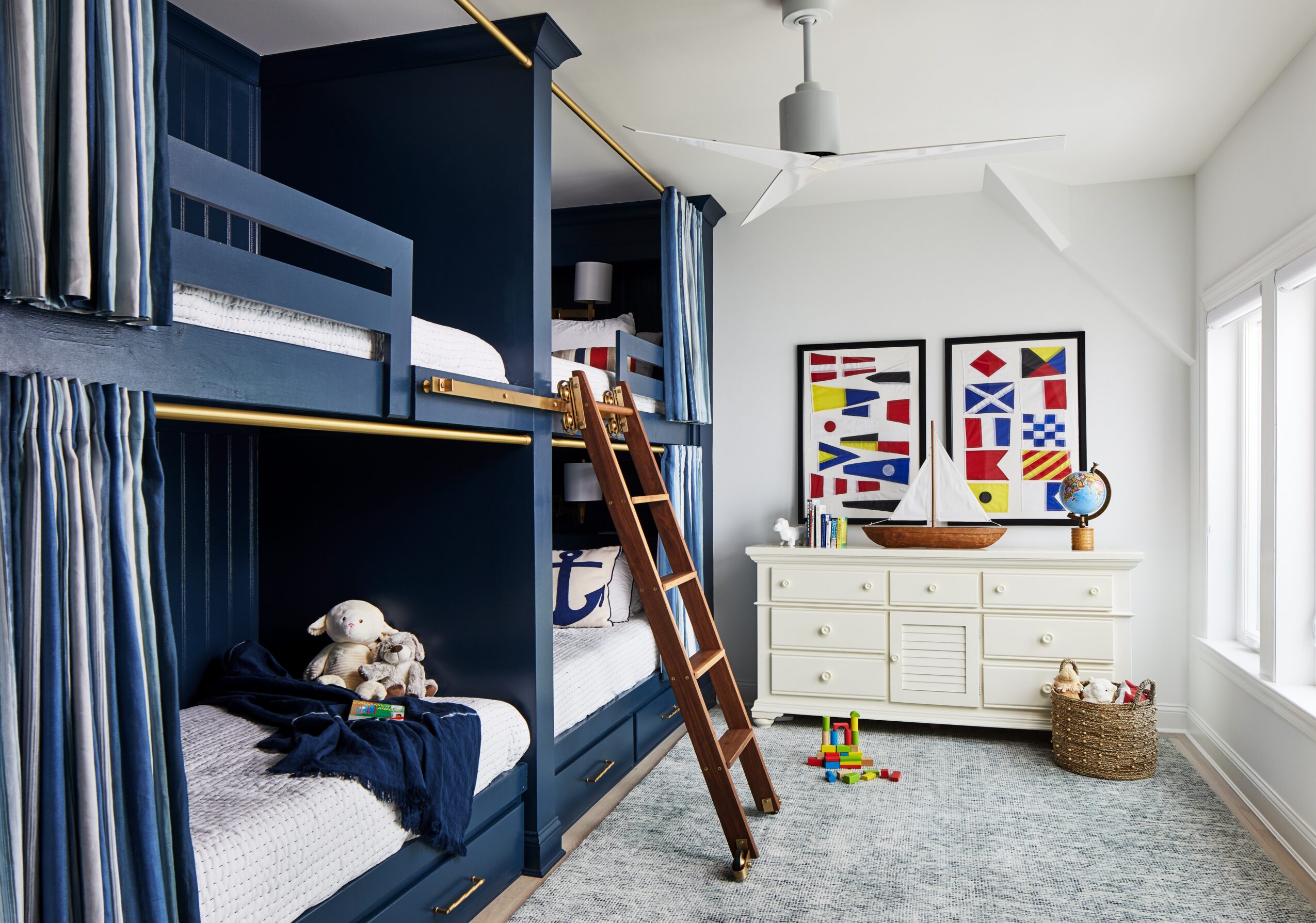 Children's bedroom with navy blue bunk beds, ladder, white dresser, colorful artwork, and toys on a light gray rug.