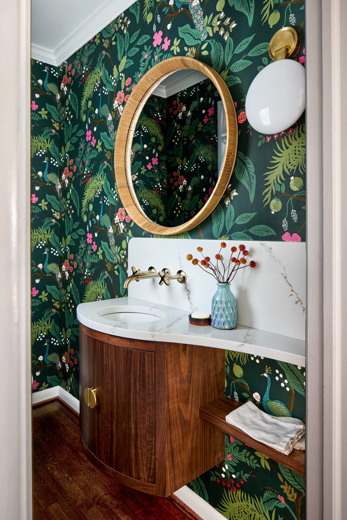 Bathroom with tropical wallpaper, round mirror, white sink, wooden cabinet, and decorative vase with red berries.