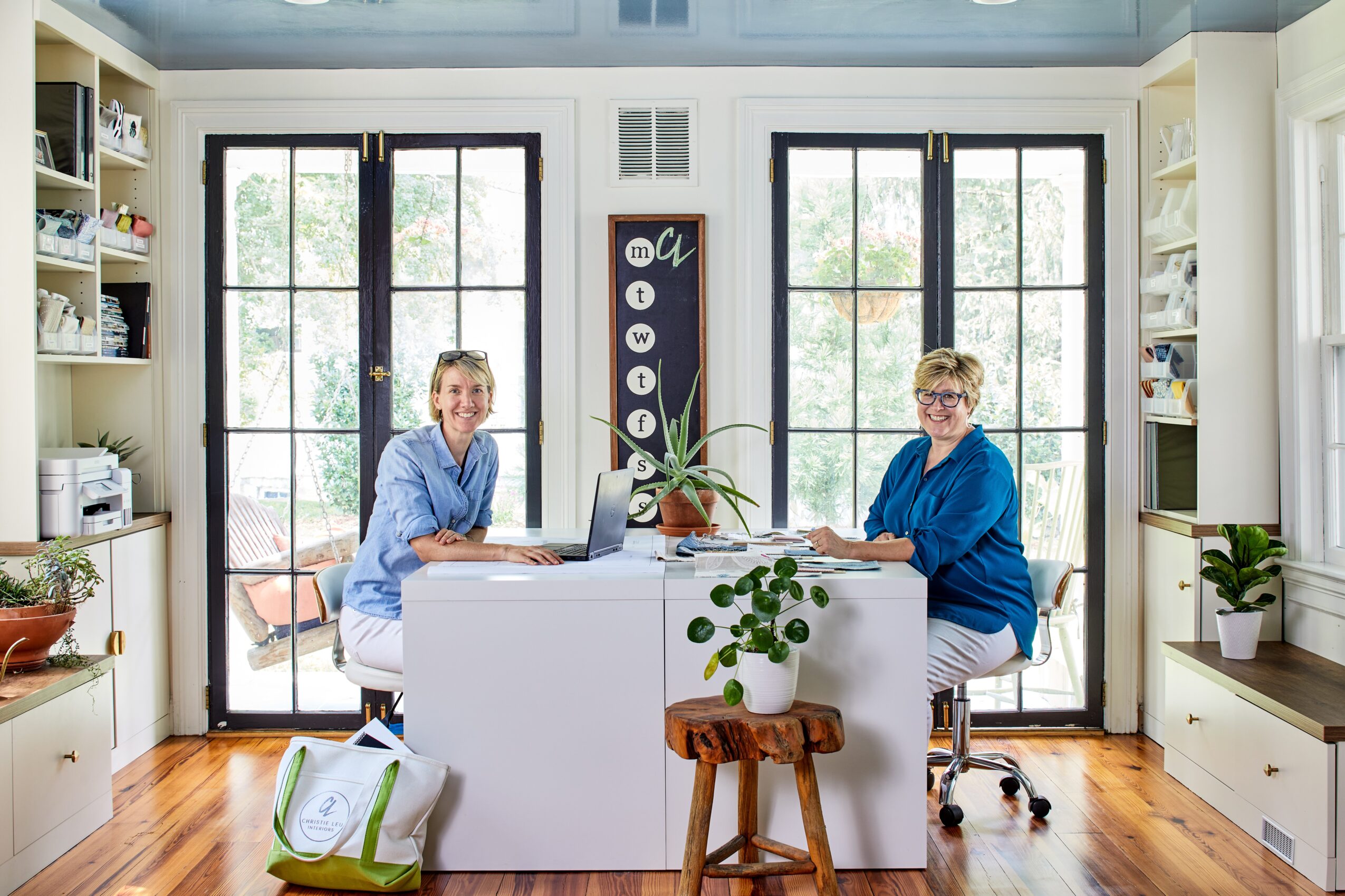 Two women sit at a white table in a bright room with large windows, bookshelves, and plants, working on laptops.
