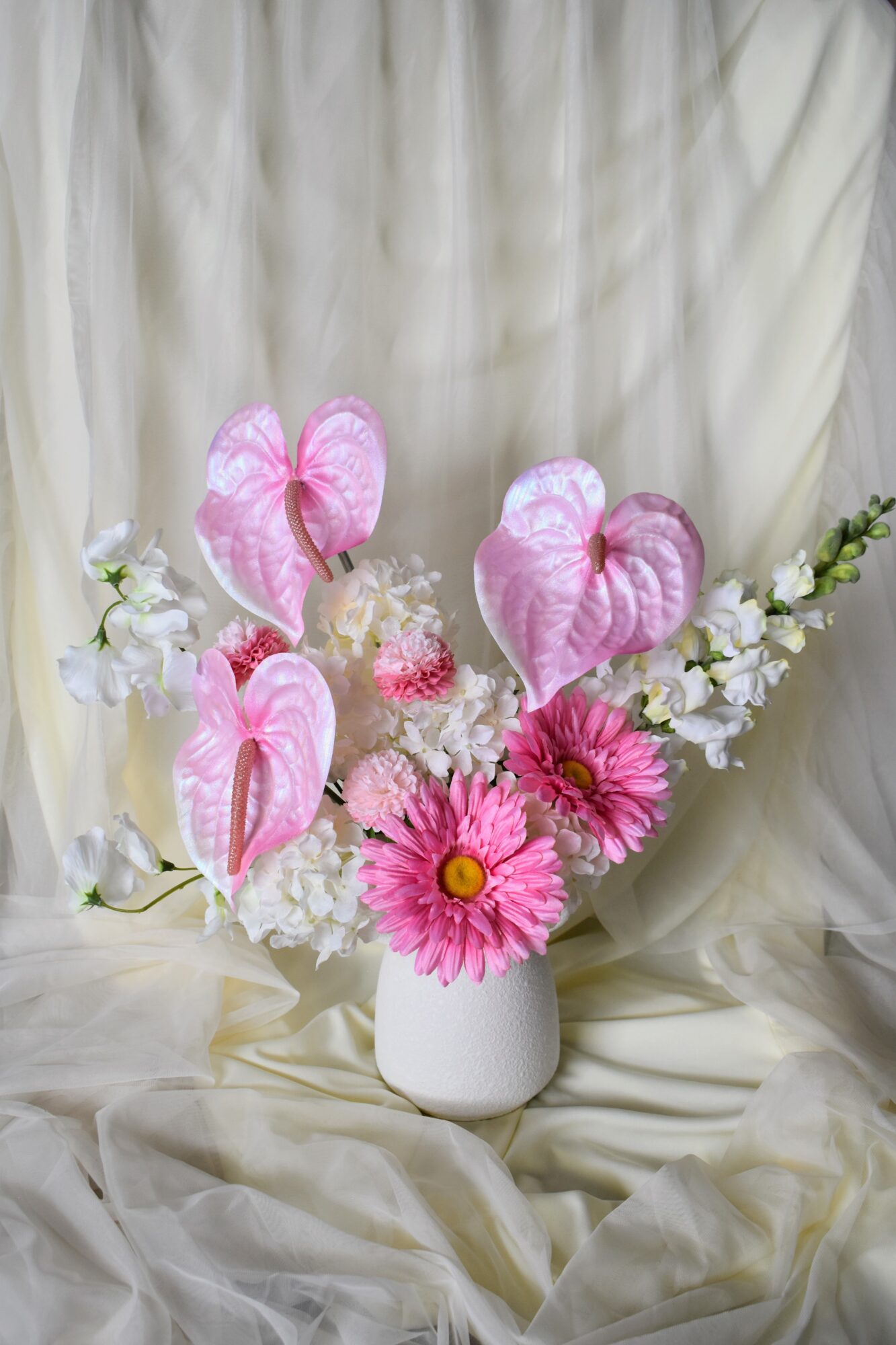 Pink and white flowers in a white vase against a cream-colored fabric background.