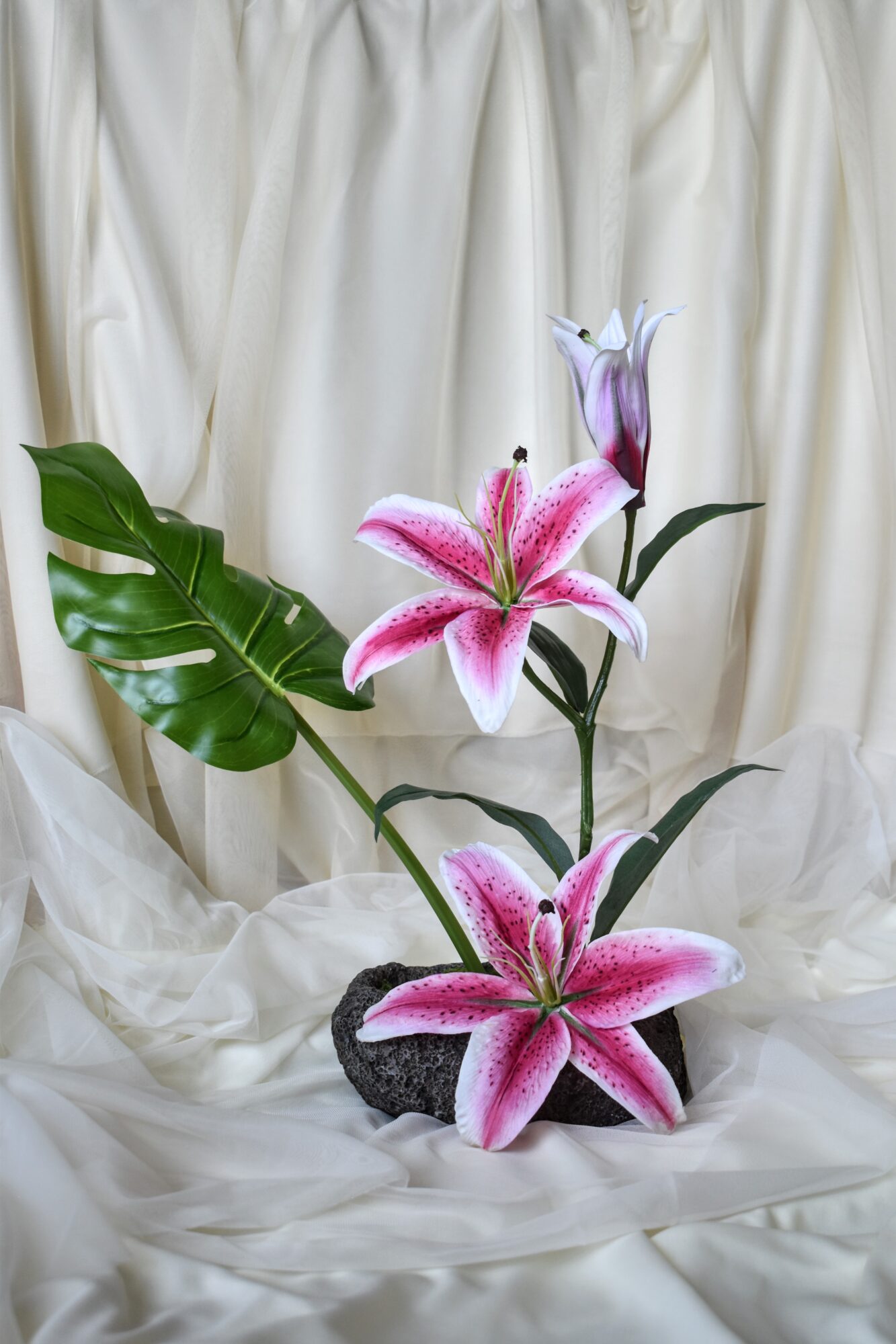 Pink and white lilies with green leaves in a pot, set against a cream-colored fabric background.