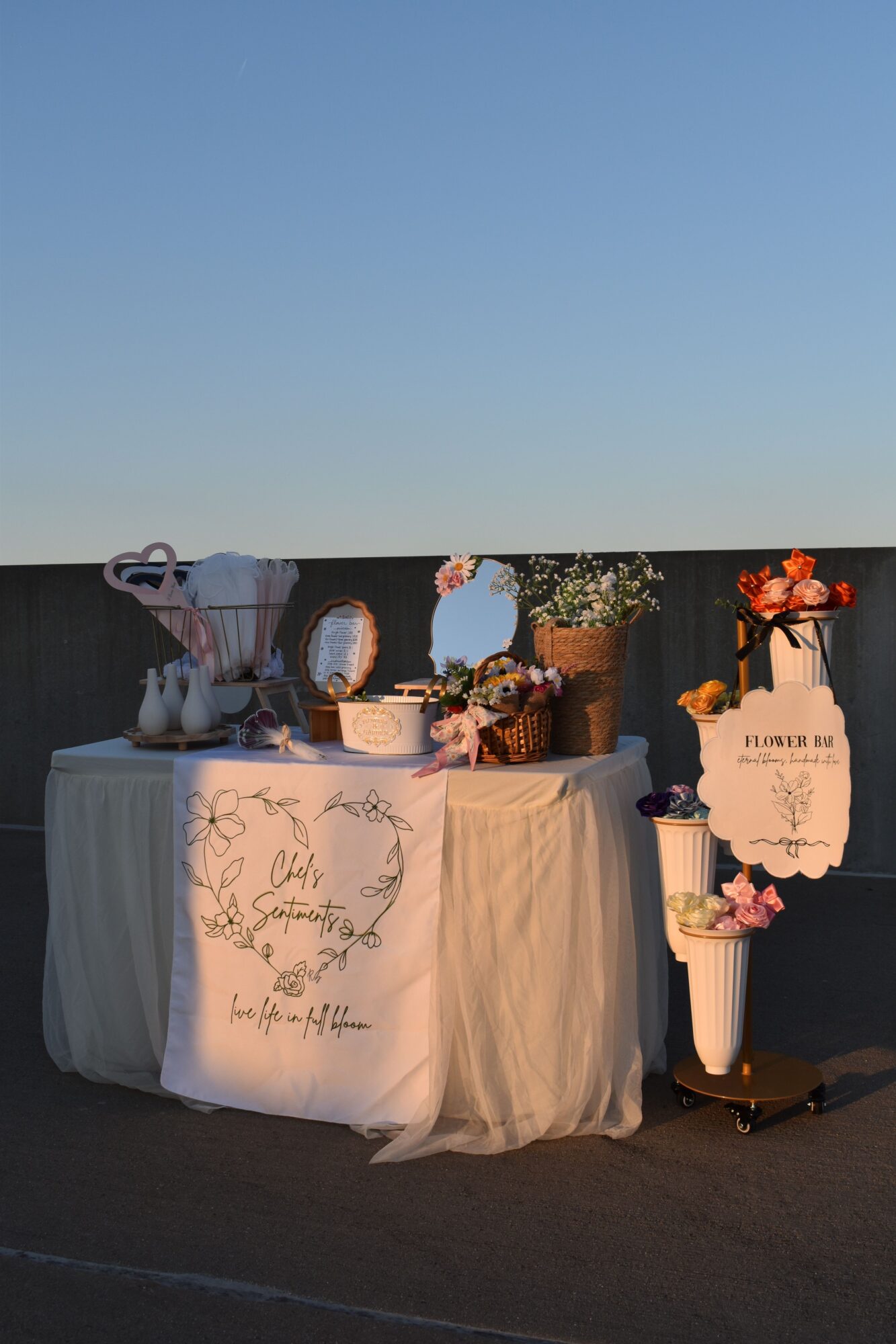 Decorative table with floral arrangements, a mirror, and a sign, set against a dark background. The table is draped with a cloth.
