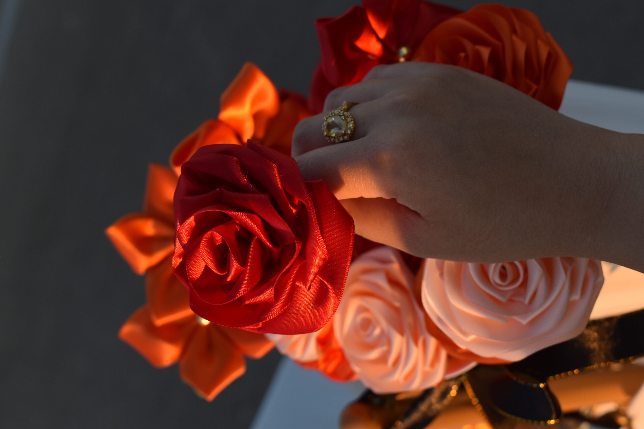 Hand holding a bouquet of red and pink roses with orange ribbons, ring visible on finger.