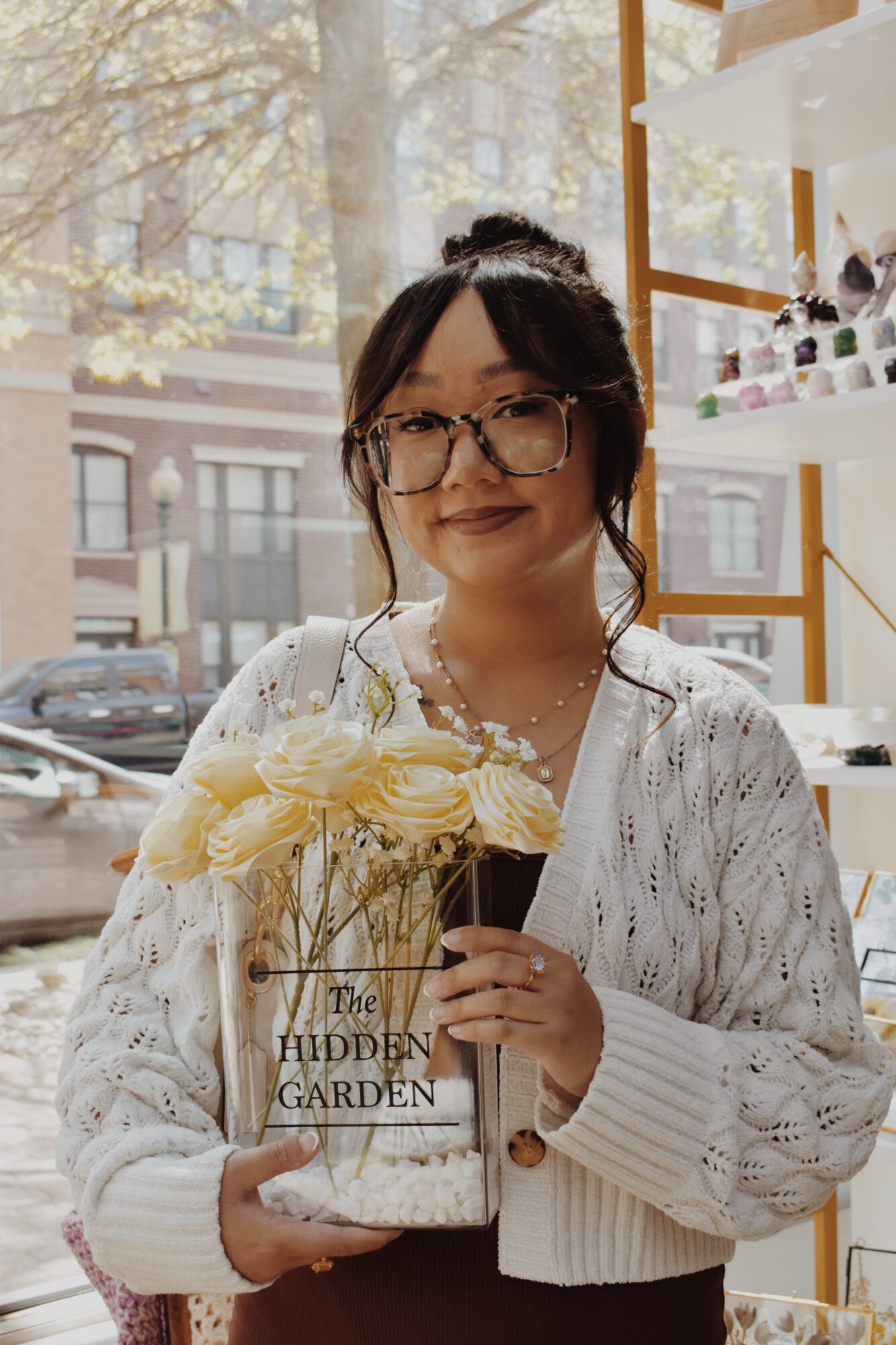 Woman with glasses holding a flower bouquet and a book in a shop with shelves, sunlight outside