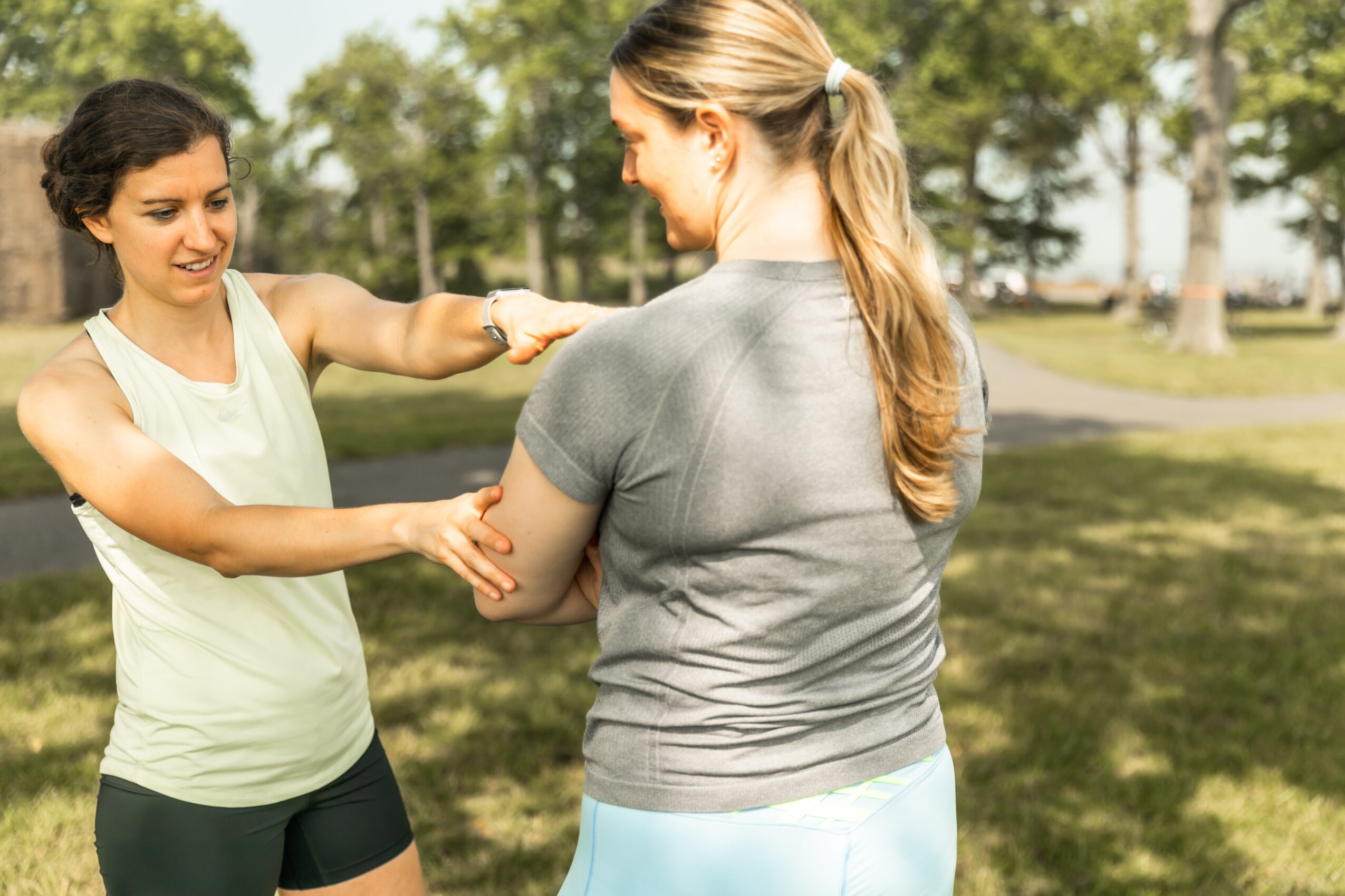 Two women outdoors, one smiling and holding the other's arm, in a park with trees and grass.