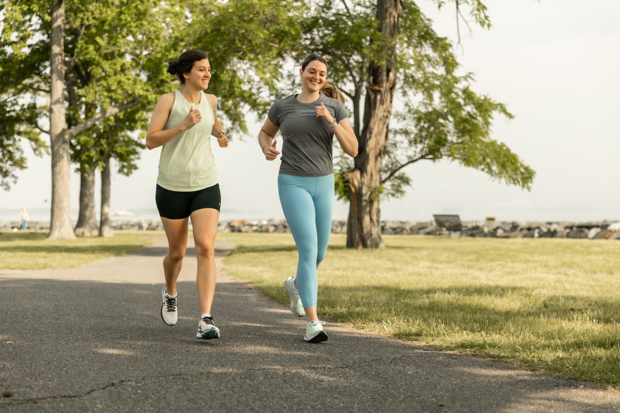 Two women jogging outdoors on a paved path with trees and a grassy area in the background.