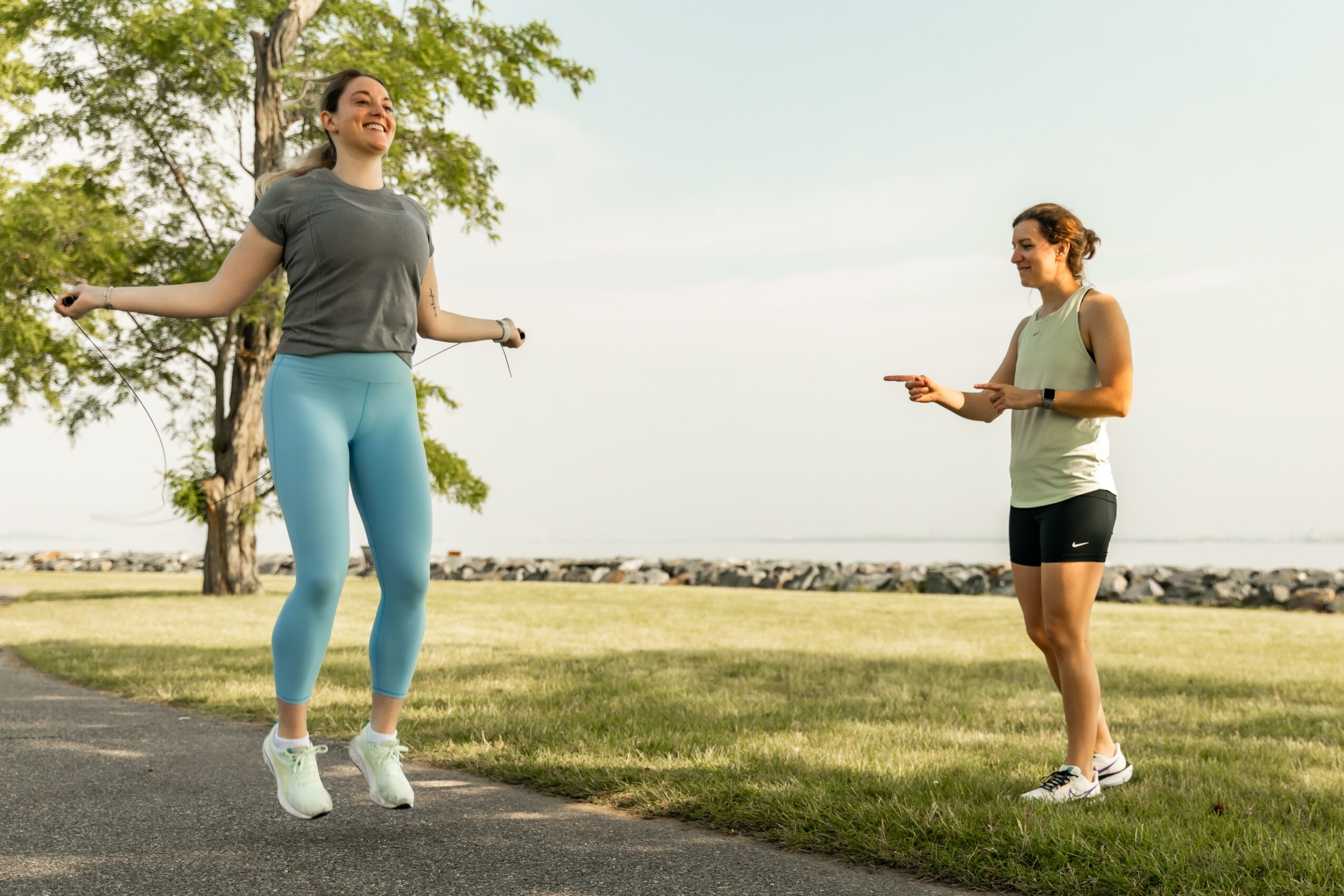 Two women outdoors near water, one jumping rope and the other holding a phone, with trees and grass in background.