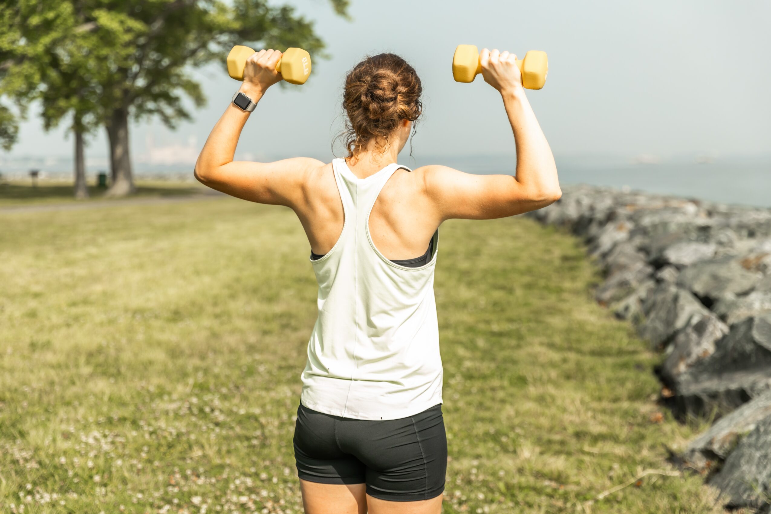 Woman lifting dumbbells outdoors near water and rocks, back view, wearing a white tank top and black shorts.