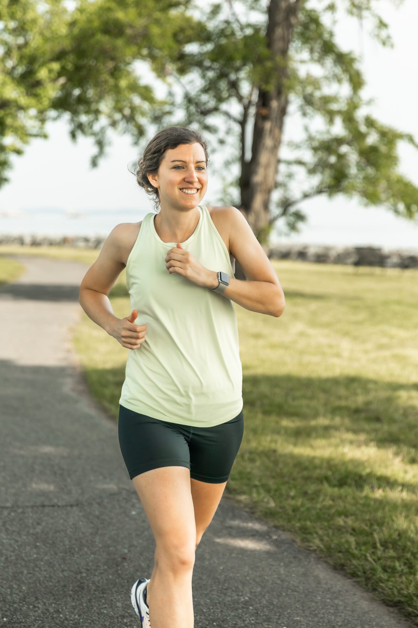 Woman jogging outdoors on a path, smiling, wearing a light tank top and black shorts, trees and grass in background.