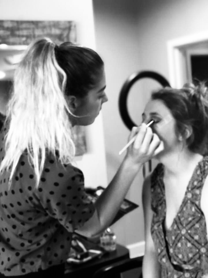 Makeup artist applies makeup to a woman sitting in a chair, in a room with a mirror and ring light, black and white.