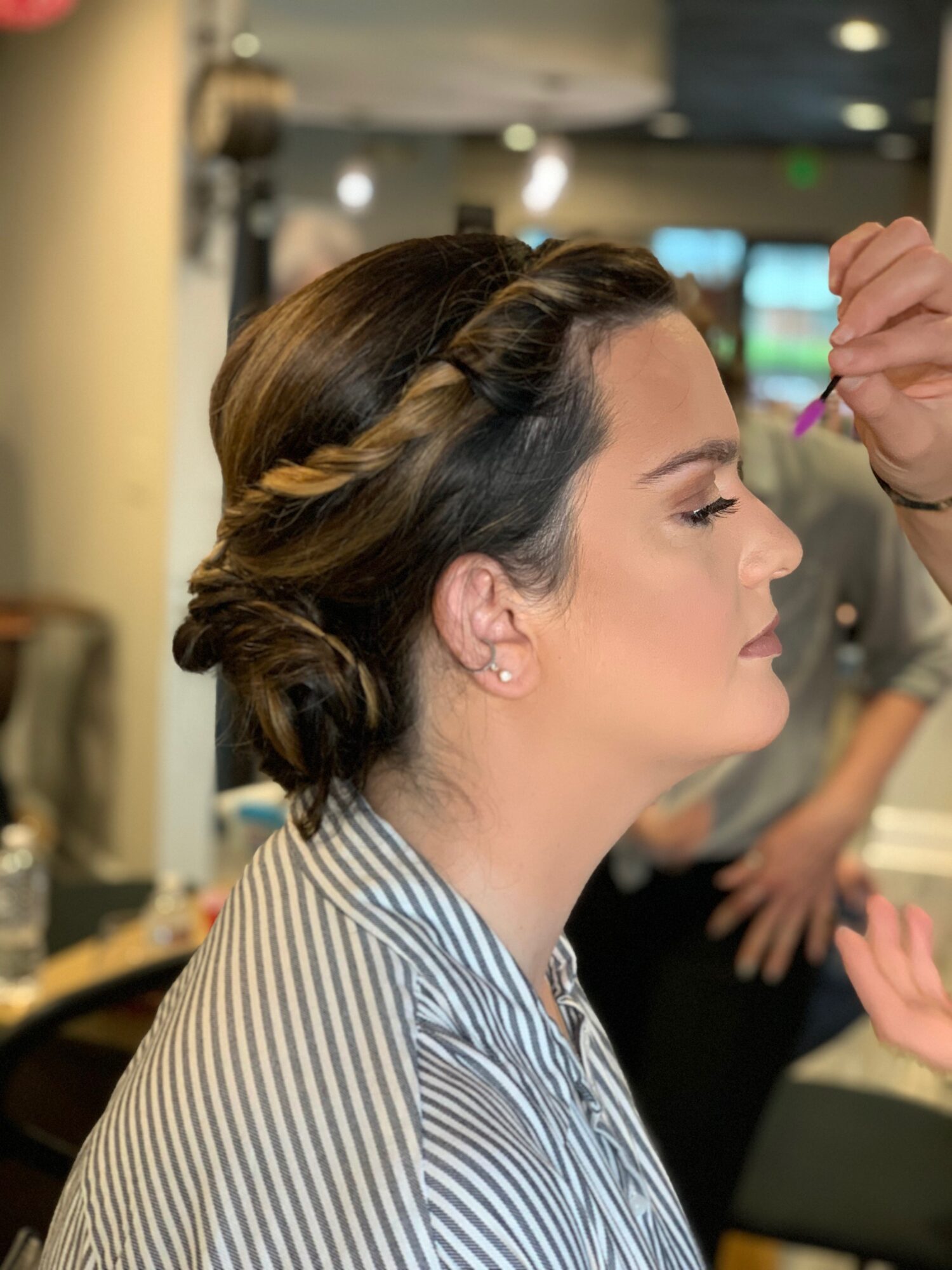 Side profile of a woman with styled hair and makeup, wearing a striped shirt, in a salon setting.