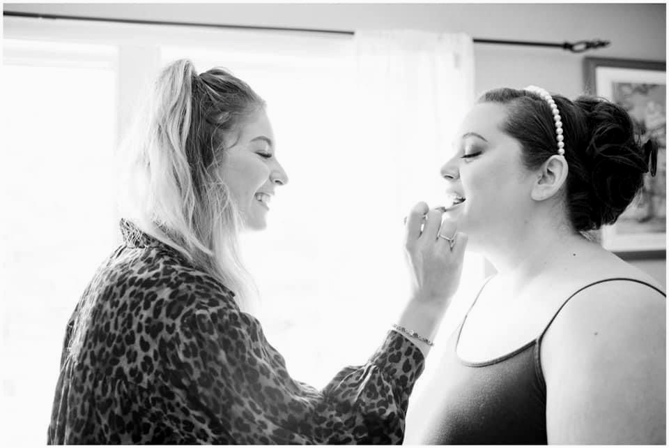 Two women smiling, one applying makeup to the other, in a bright room with curtains and framed pictures, black and white.