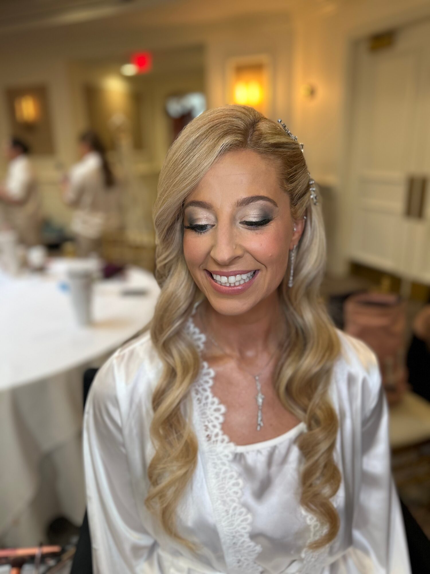 Smiling woman with long wavy blonde hair in a white satin robe, in a decorated indoor setting.