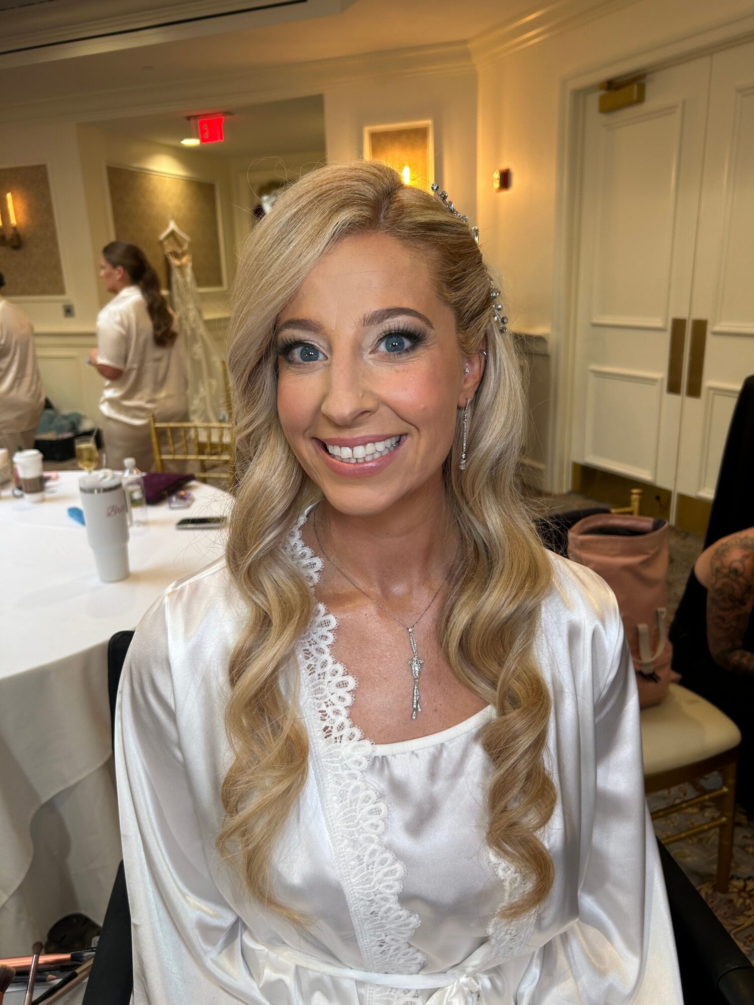 Woman with long blonde hair smiling, wearing a white blouse, in a decorated indoor setting.