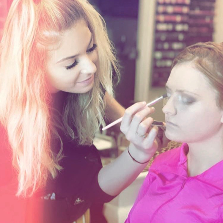Makeup artist applies makeup to a young woman with red hair, wearing a pink shirt, in a studio setting.