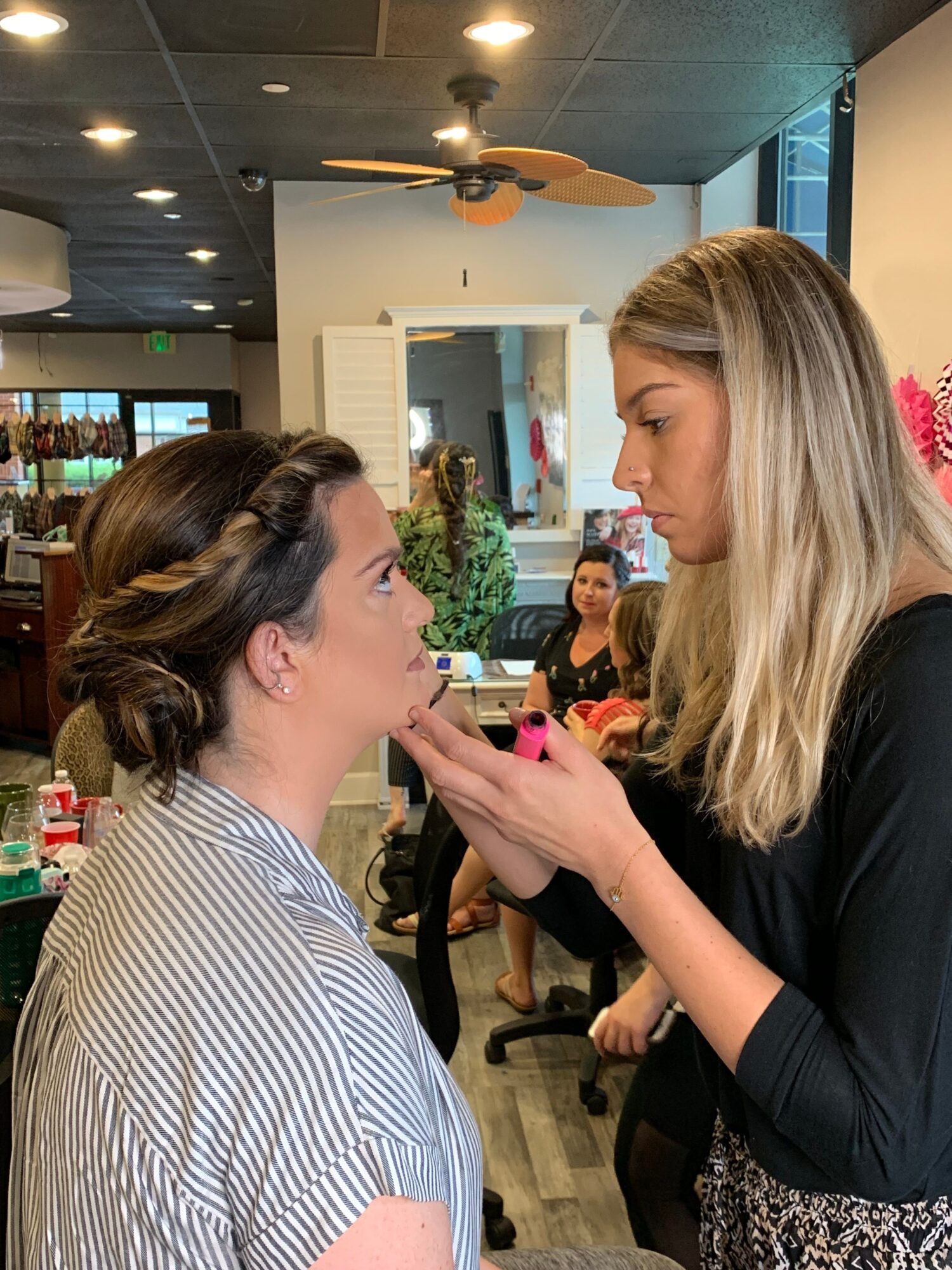 Two women face each other indoors, one applying makeup or lipstick to the other, with a background of a hallway and plants.