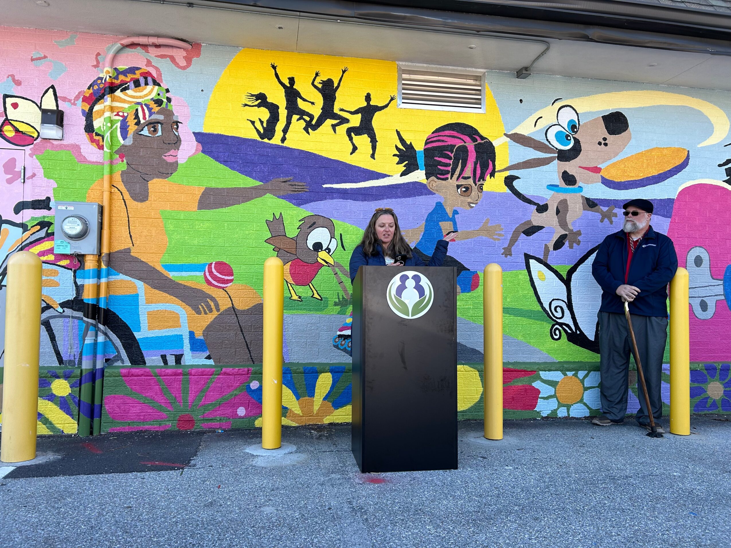 Colorful mural with diverse characters, animals, and abstract designs, featuring a woman speaking at a podium and a man standing nearby.