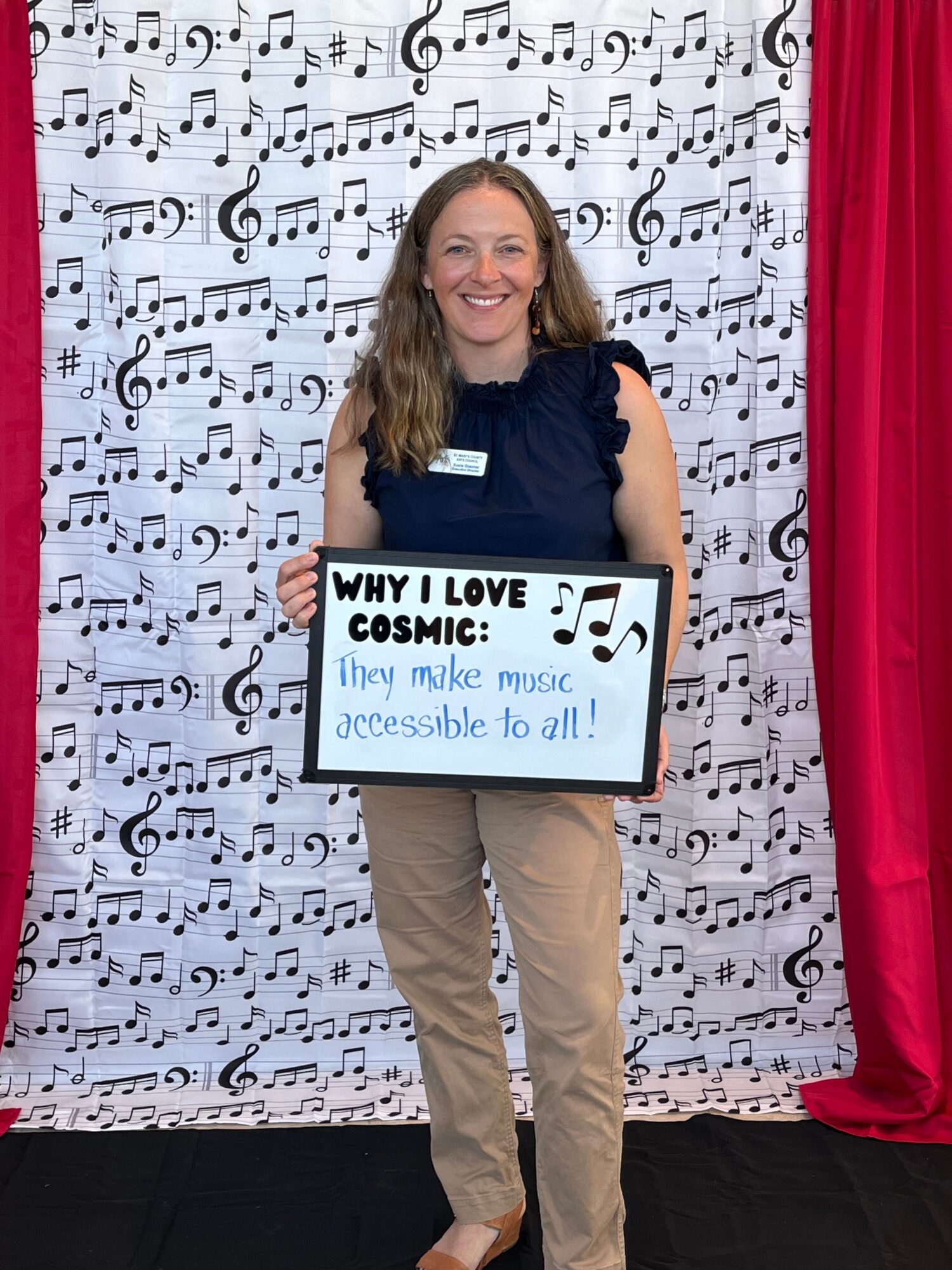 Person holding a sign with text, standing in front of a music-themed backdrop, smiling, wearing beige pants and a dark top.
