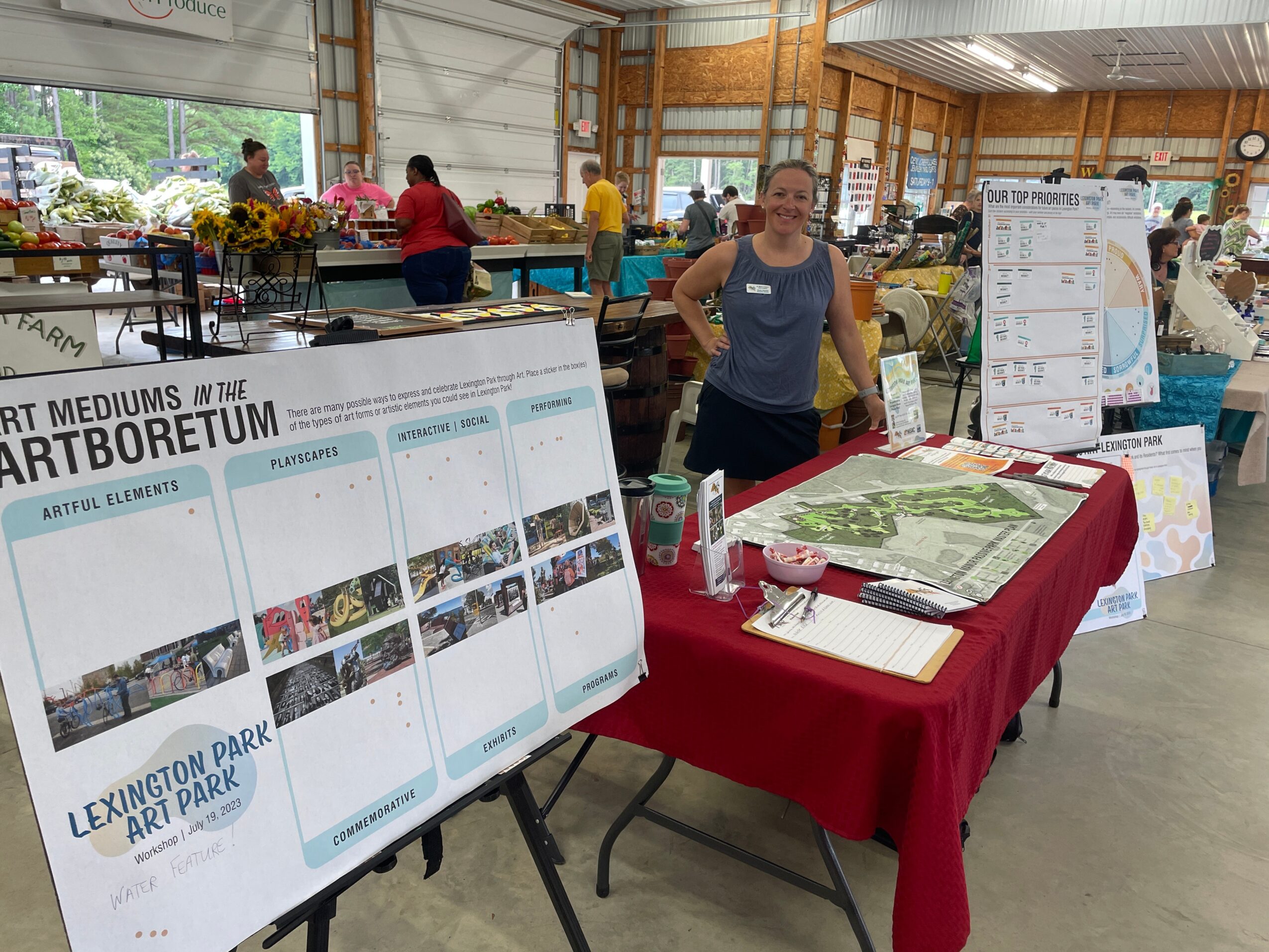 Woman standing behind a table with a large map and informational displays at an indoor event, with other booths and people in the background.